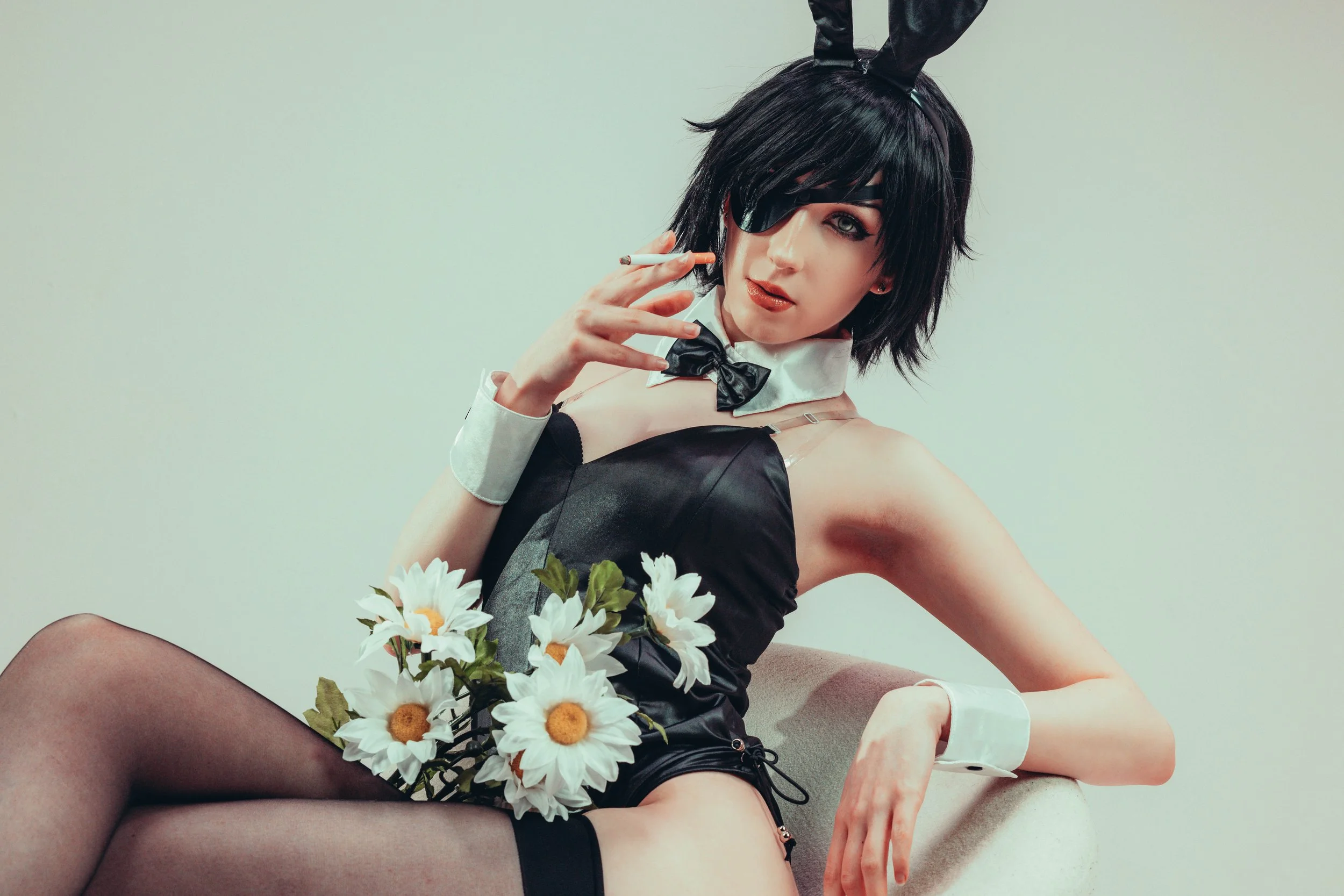 Woman with black hair, eyepatch, bunny ears, wearing black lingerie, holding flowers, smoking, sitting on a beige object against a plain background.