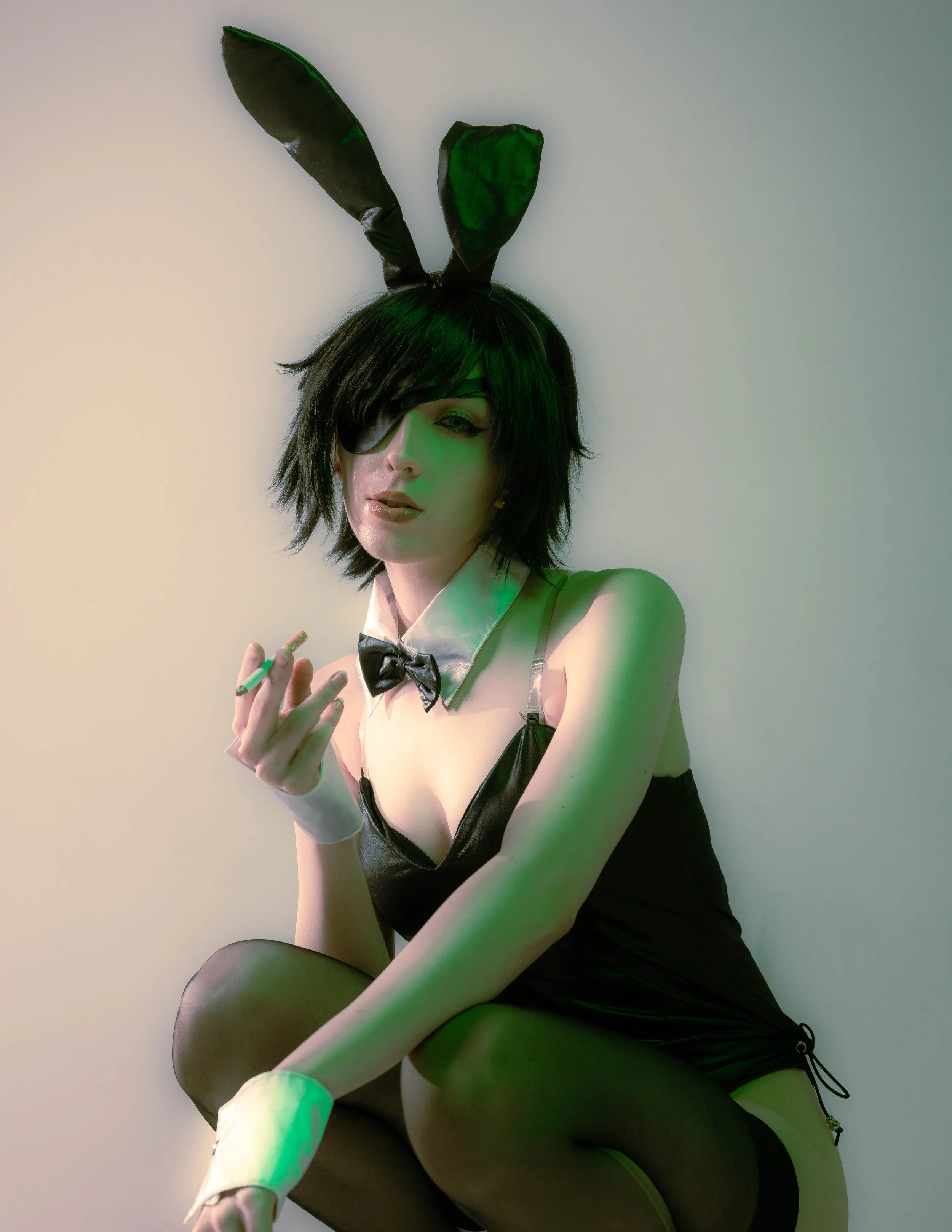 A woman with black hair, wearing an eye patch, bunny ears headband, a black bunny costume, and thigh-high stockings, sitting on the floor against a plain wall in a costume.