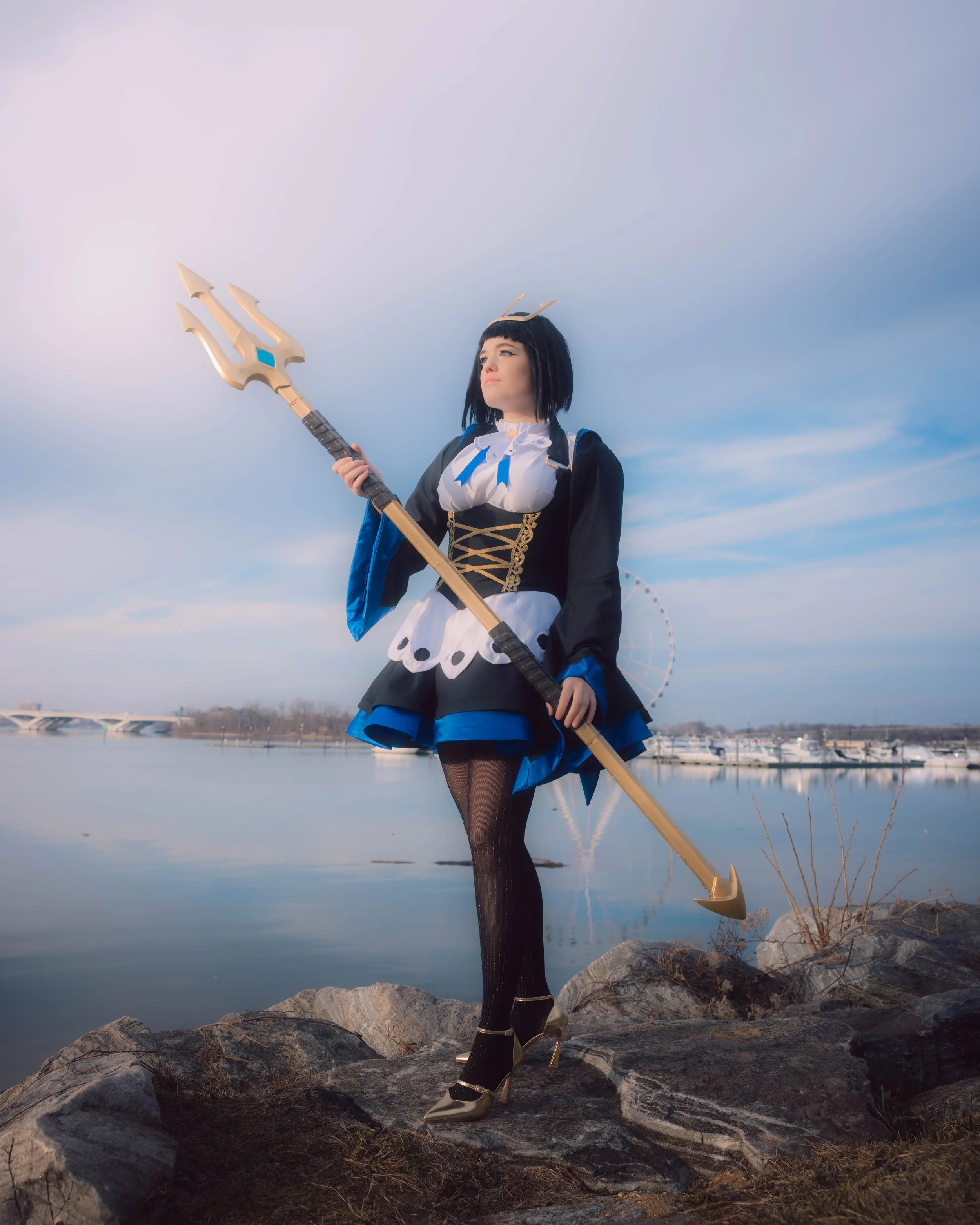 A woman dressed as a fantasy character stands on rocks near water, holding a trident, with a parasol and bridge in the distance.