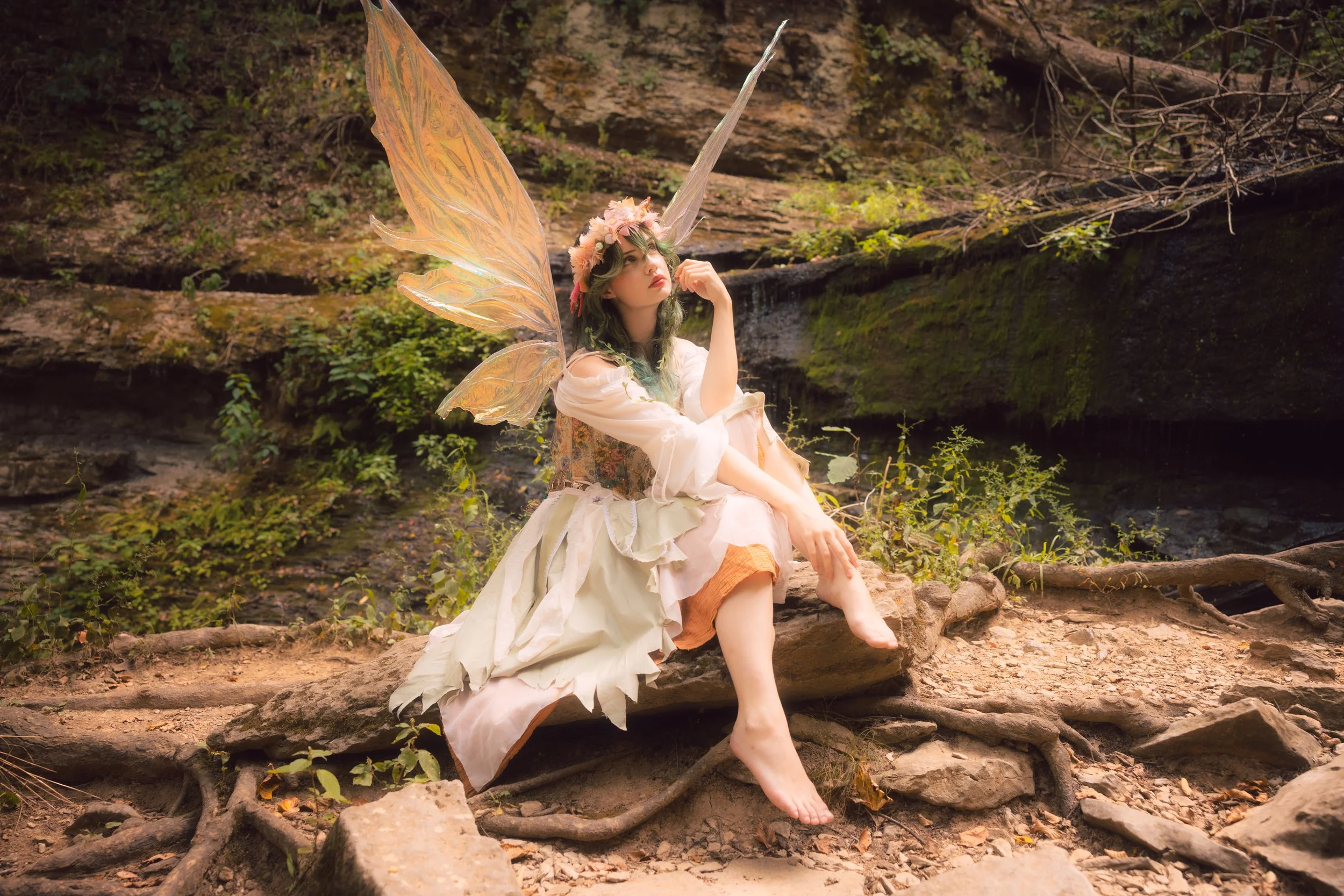 A person dressed as a fairy with large, translucent, orange wings and a flower crown sitting on rocks near a waterfall in a wooded area.