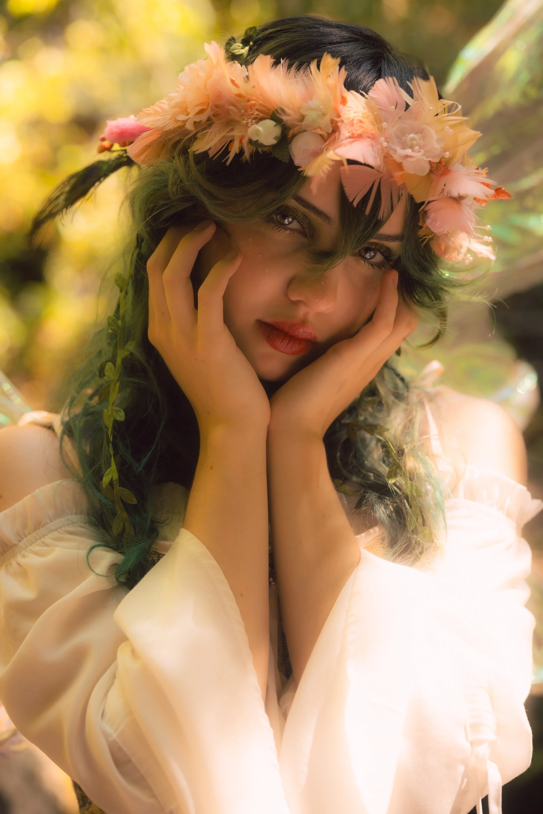 A young woman with dark hair styled in loose waves, wearing a floral crown made of pink and peach feathers and flowers, looking directly at the camera with a soft expression, outdoors with a blurred background of greenery and sunlight.