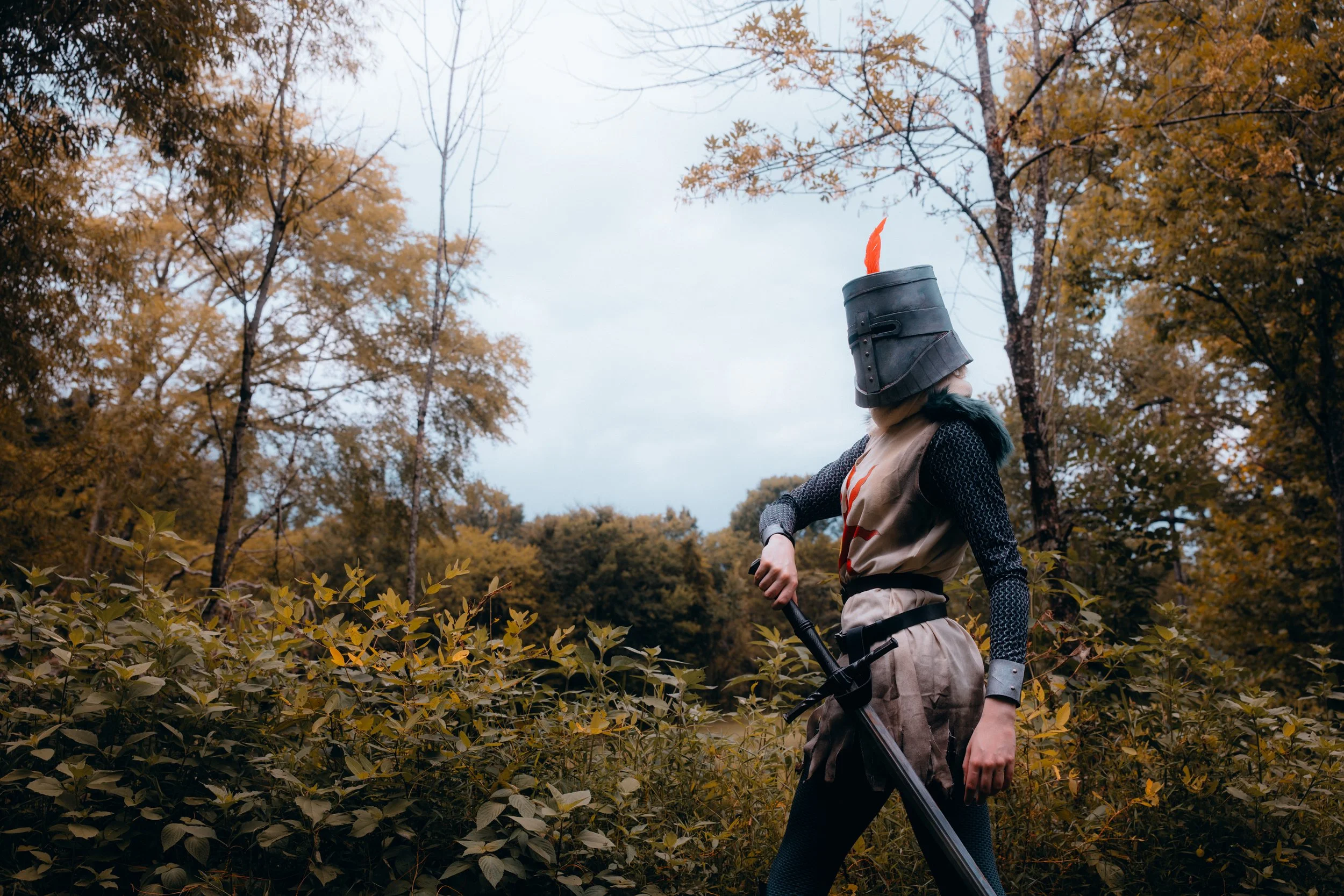 A person dressed as a medieval knight, wearing a helmet with a red feather, walking through a forest with tall trees and yellow fall foliage.