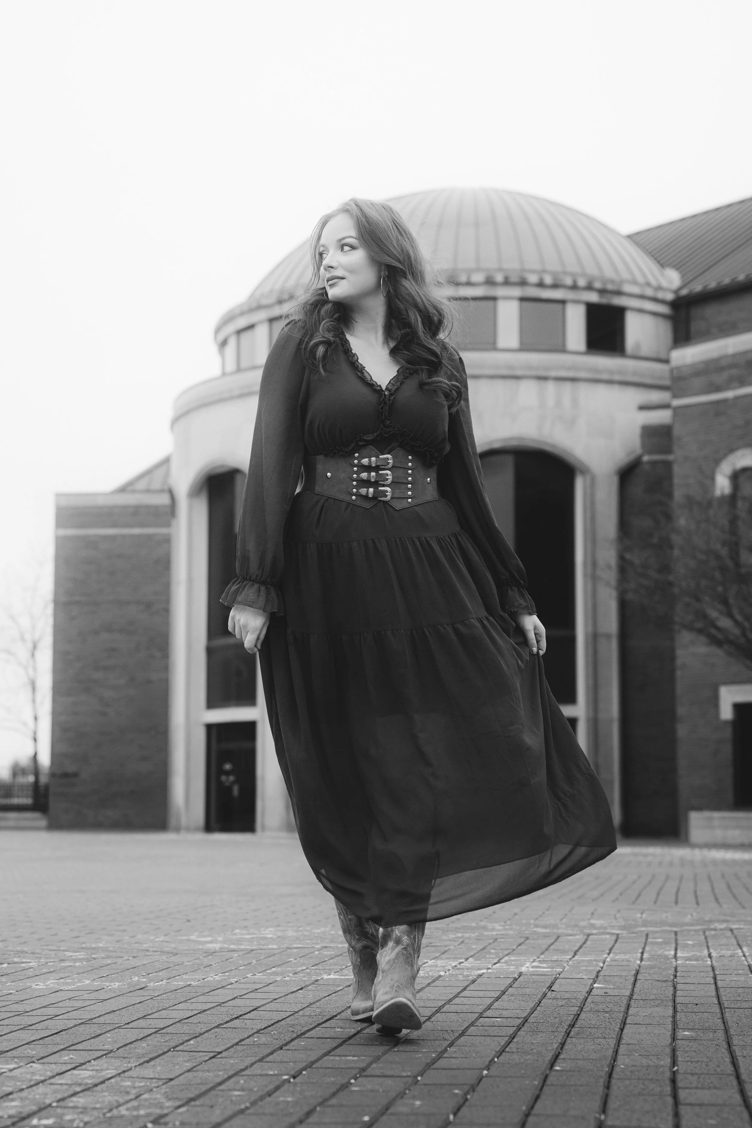 A woman with long wavy hair wearing a long, flowing dress with a wide belt, walking outdoors on a paved area in front of a building with a rounded roof, captured in black and white.