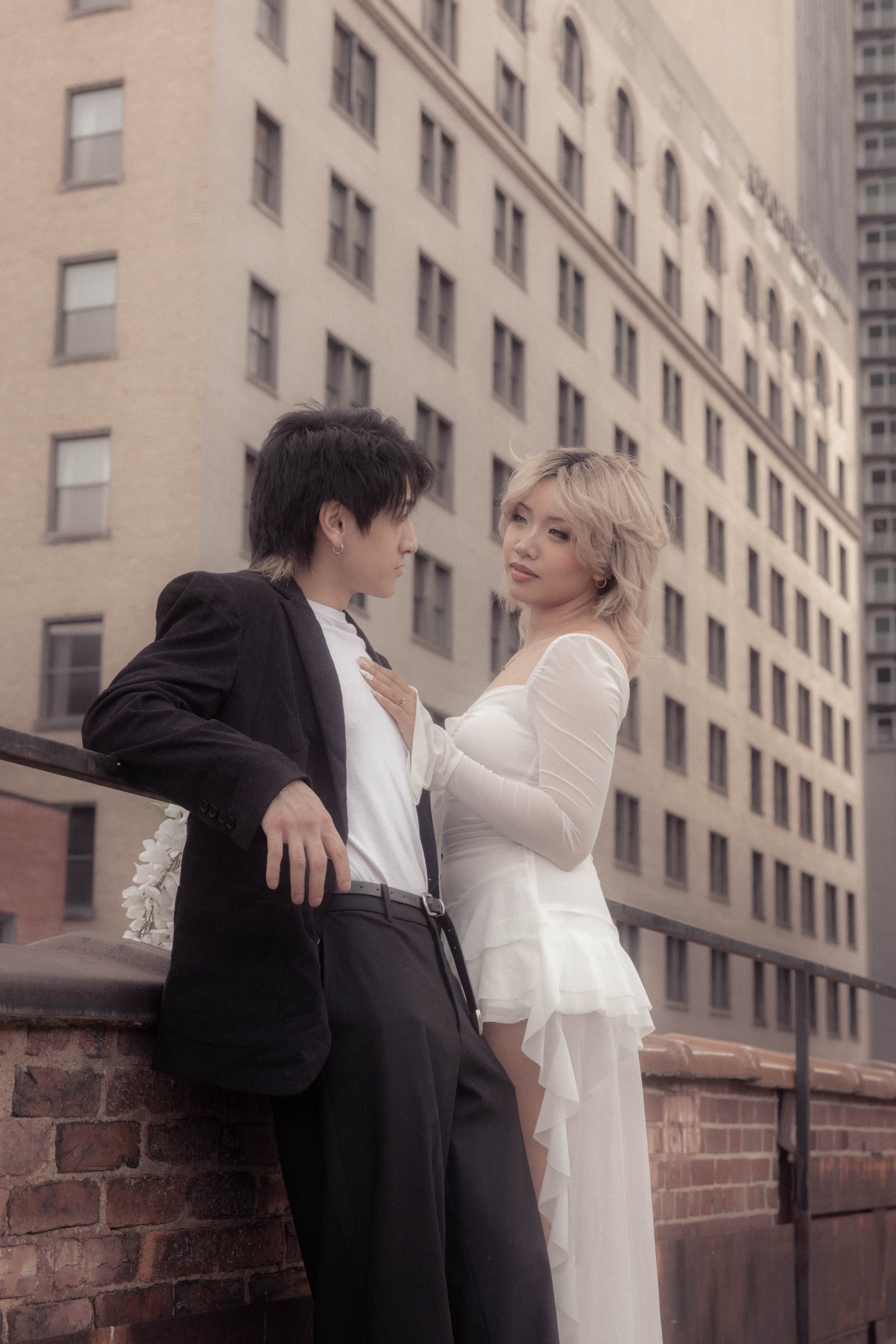 A man in a black suit and a woman in a white dress on a rooftop in an urban setting with tall buildings.