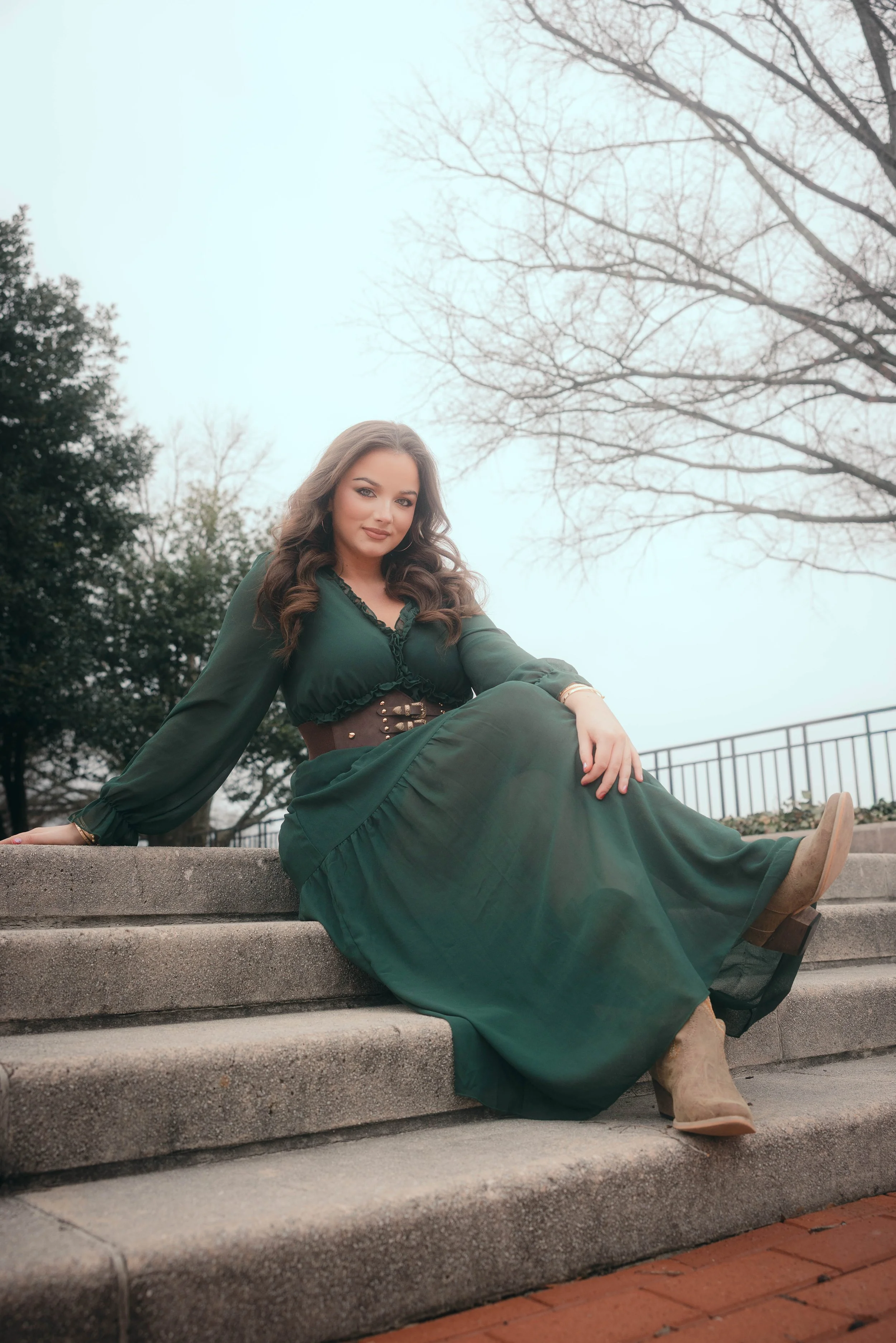 A woman with long brown hair sitting on outdoor concrete steps, wearing a green dress, beige boots, and a brown belt, with trees and a railing in the background.