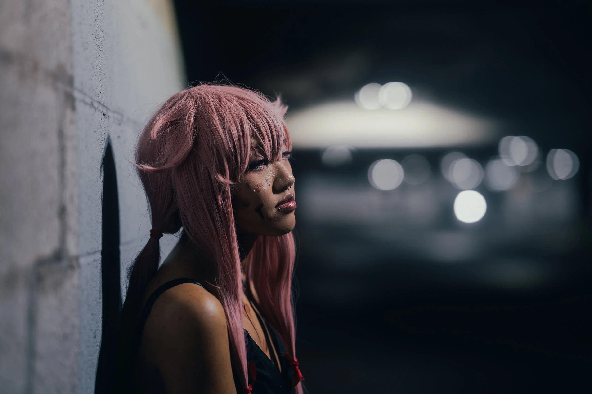 A woman with pink hair and facial piercings leaning against a concrete wall in a dimly lit urban setting.