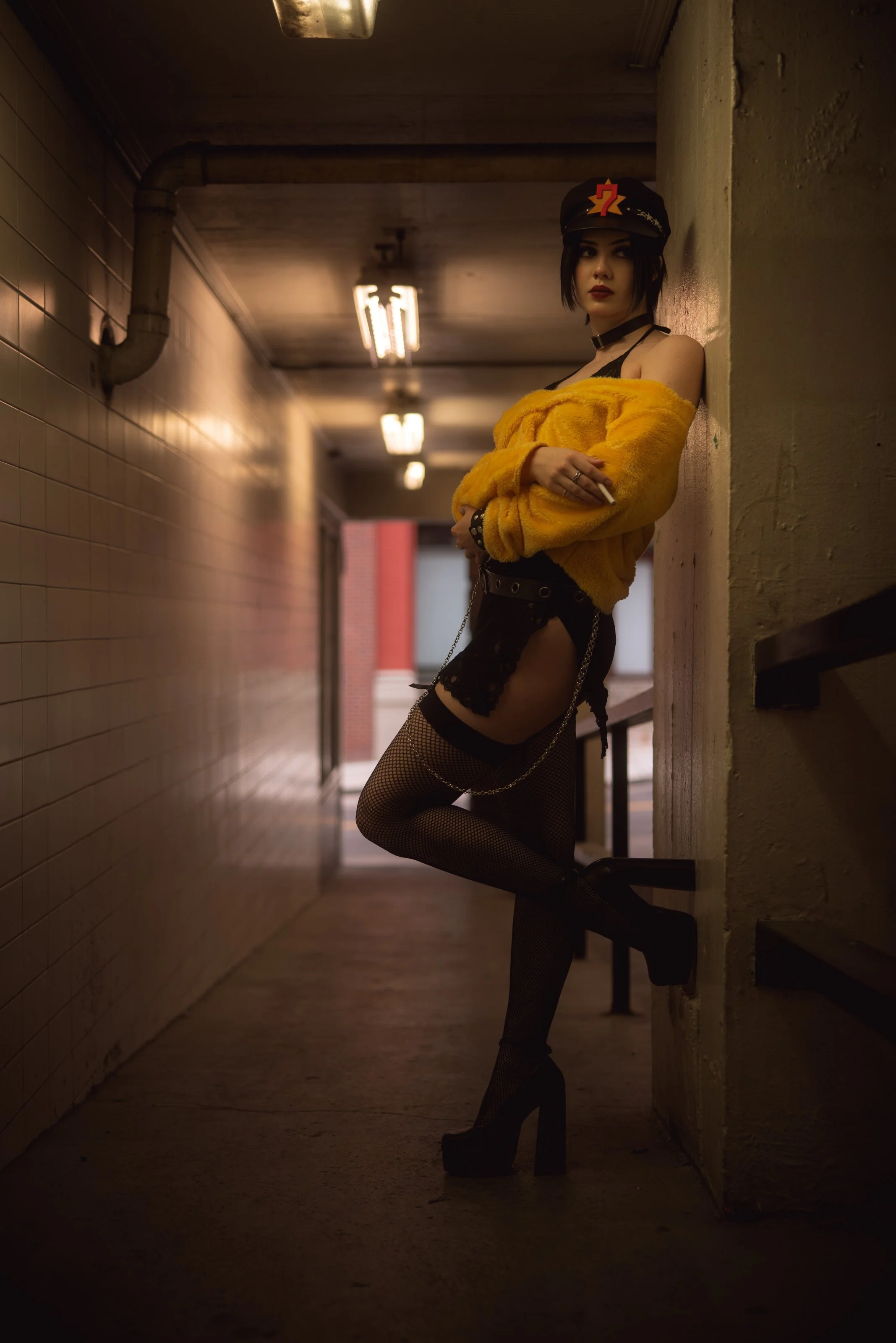 A woman with dark hair and dark makeup standing in a dimly lit corridor, leaning against a wall. She is wearing a black hat with a red question mark pin, a yellow furry jacket, fishnet stockings, and high-heeled boots. The corridor has a tiled wall o