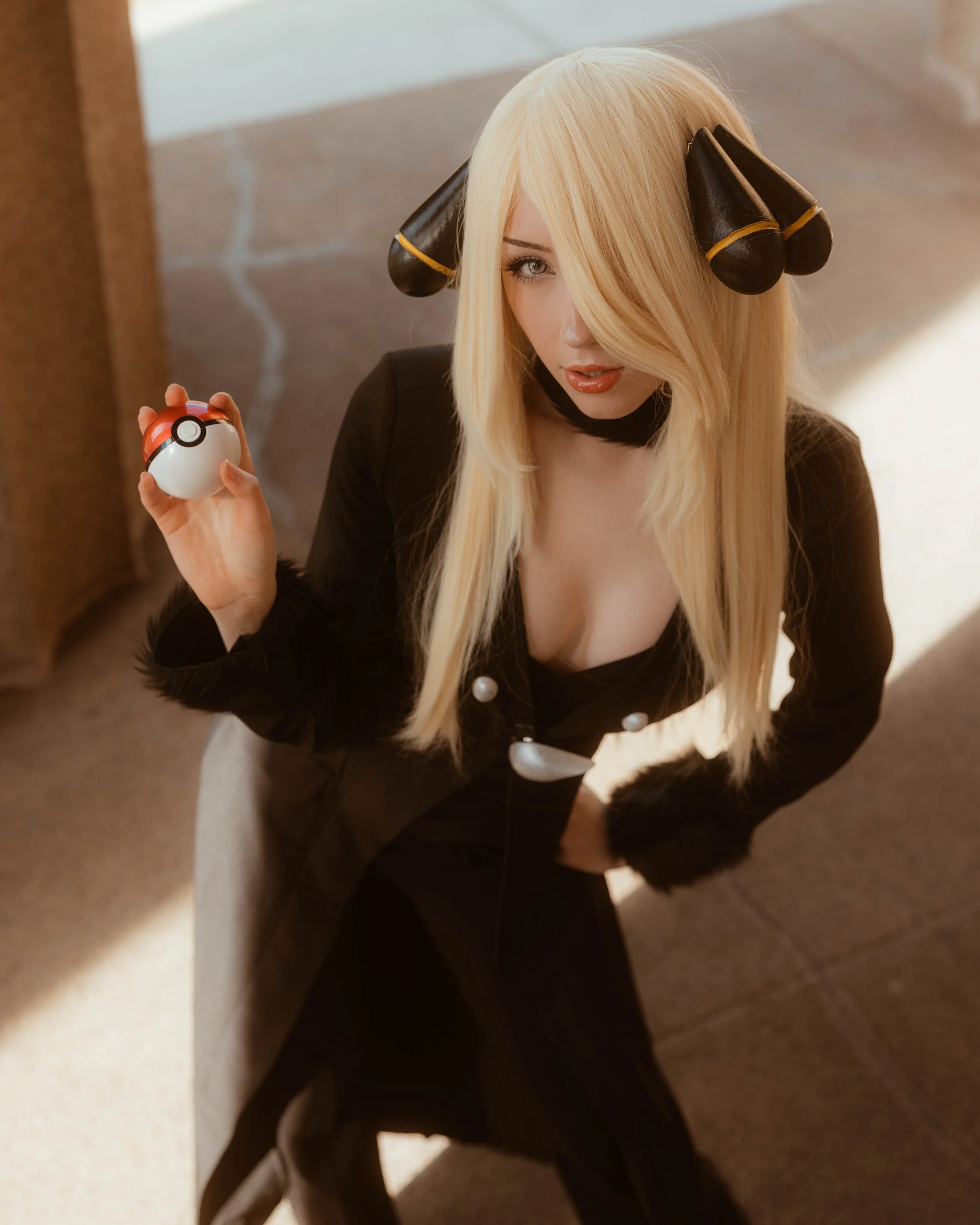 A woman with long blonde hair, wearing a black blazer with pearl buttons, sitting on a staircase. She has headphones resembling Pokemon's Lullaby, holding a Poké Ball in her hand, and looking at the camera with a slight smile.