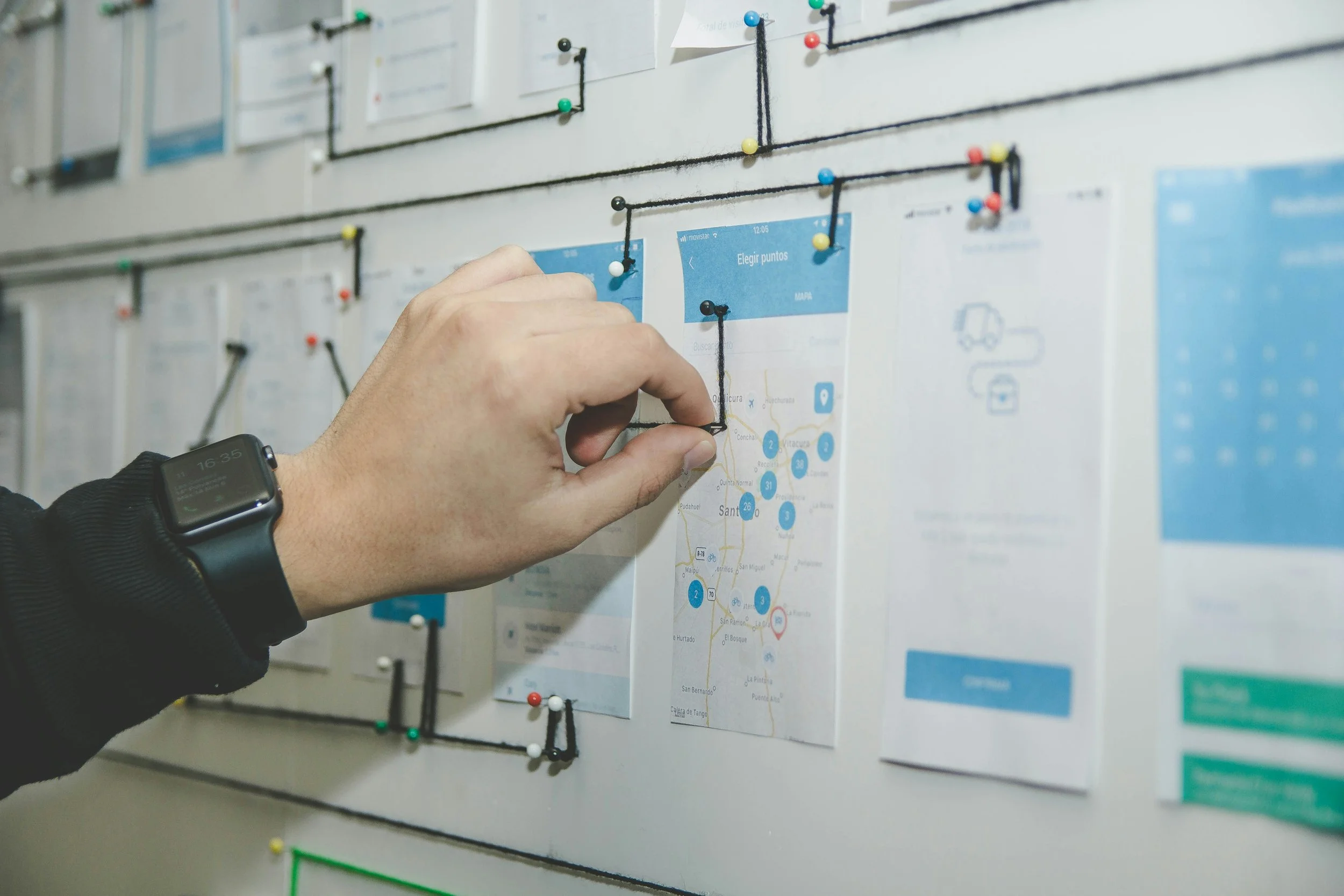 A person with a smartwatch on their wrist pins a black string connection between yellow and white push pins on a whiteboard covered with maps, text, and diagrams connected with string and push pins.