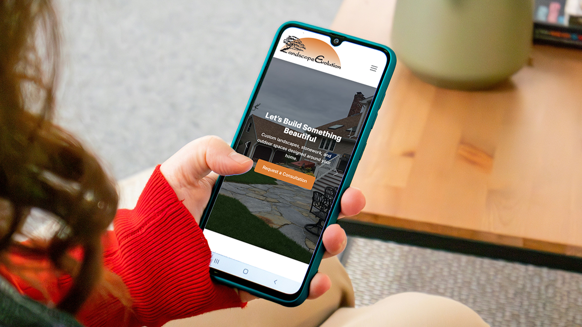 Person holding a smartphone displaying the Landscape Evolution landscaping website with a button that says "Request a Consultation" in a cozy living room.