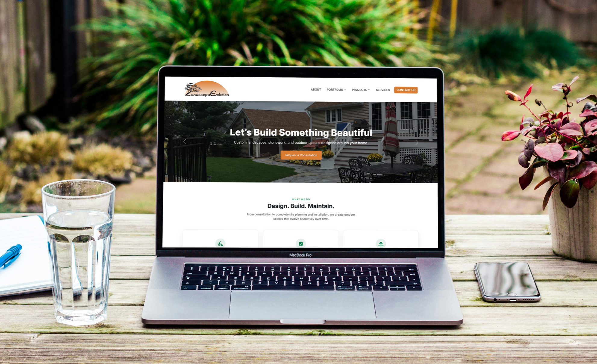 A silver MacBook Pro laptop displaying a landscaping company's website, placed on a wooden outdoor table. To the left, there's a glass of water with bubbles, a blue pen, and a notebook. To the right, a smartphone lies on the table. In the background,