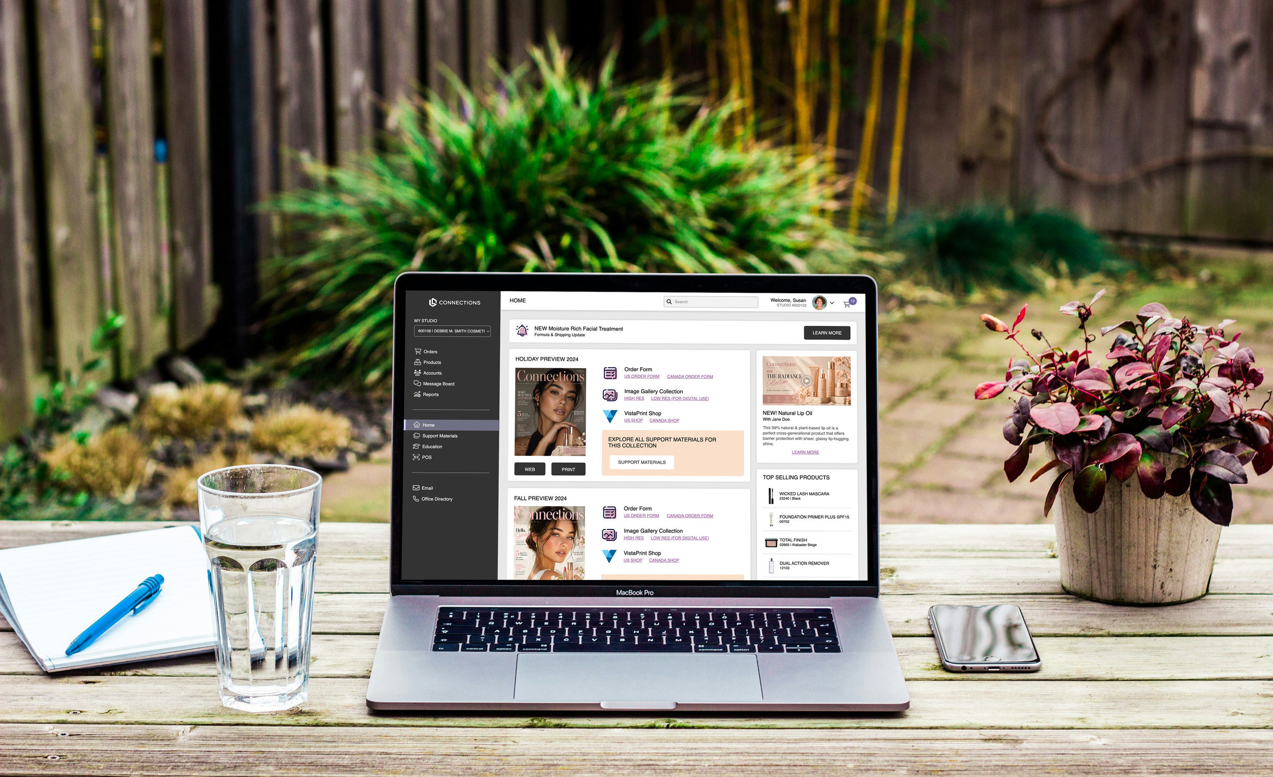 A laptop open on a wooden outdoor table displaying a website for a skincare and beauty store, with a glass of water, a notebook with a pen, and a smartphone nearby. In the background, there are green plants and a wooden fence.