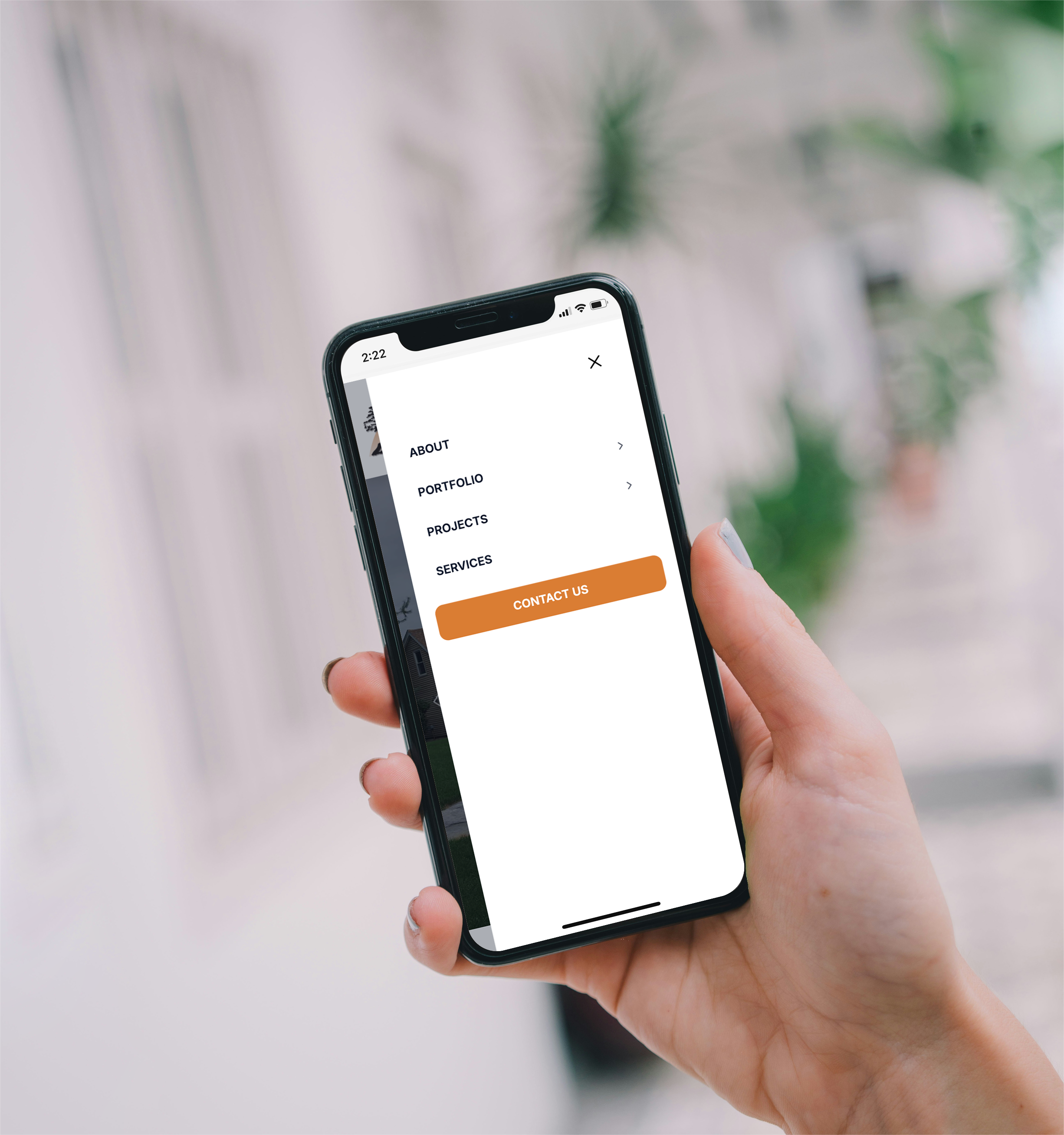 Close-up of a person's hand holding a smartphone with a visible screen displaying a website navigation menu with options including About, Portfolio, Projects, Services, and Contact Us, against a blurred background of green foliage.