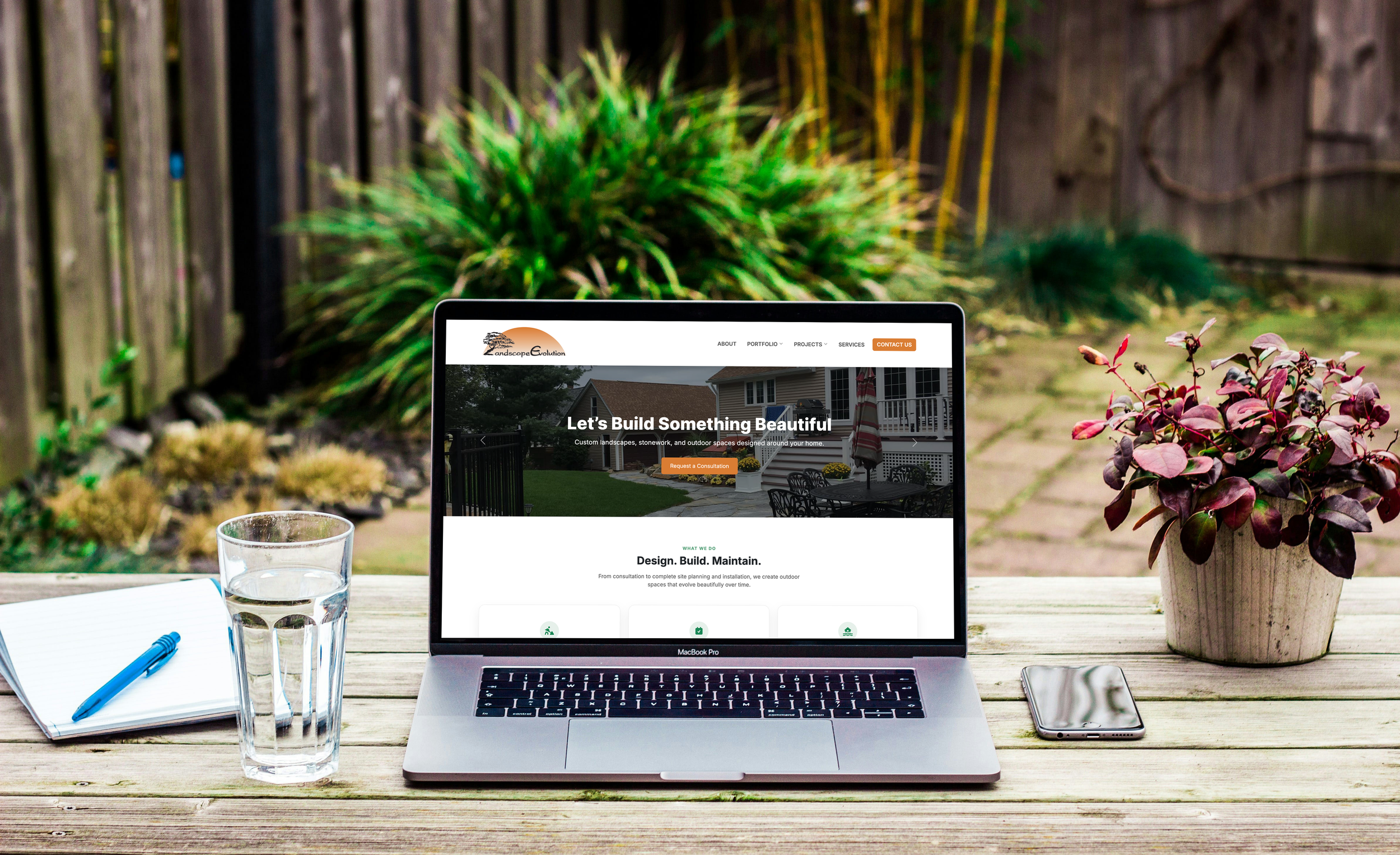 An outdoor workspace with a MacBook Pro on a wooden table, displaying a landscaping company's website. A glass of water, a notebook with a blue pen, and a smartphone are also on the table. In the background, there is greenery and a wooden fence.
