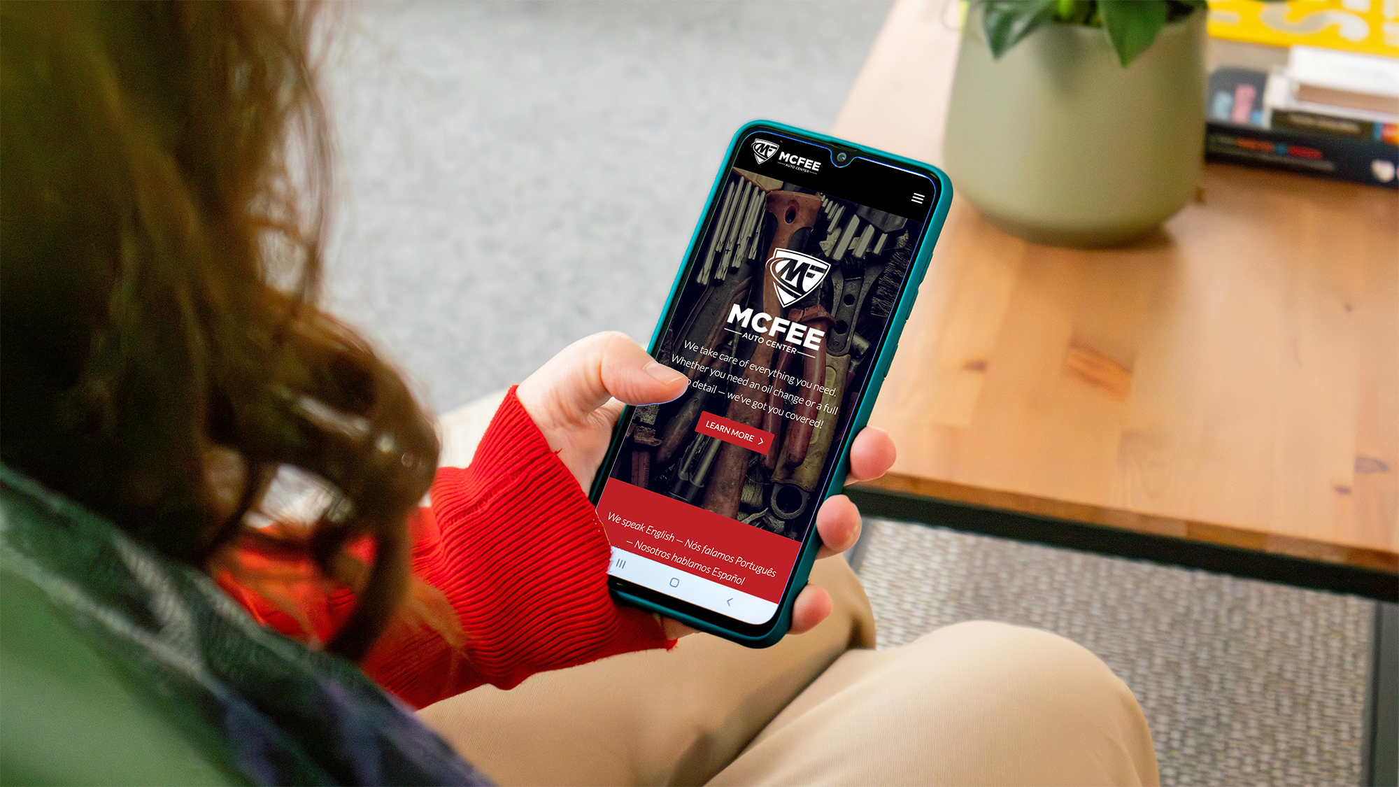 A woman wearing a red sweater is holding a smartphone displaying the MCREE Auto Center website. The website features tools and equipment in the background and offers services in multiple languages.