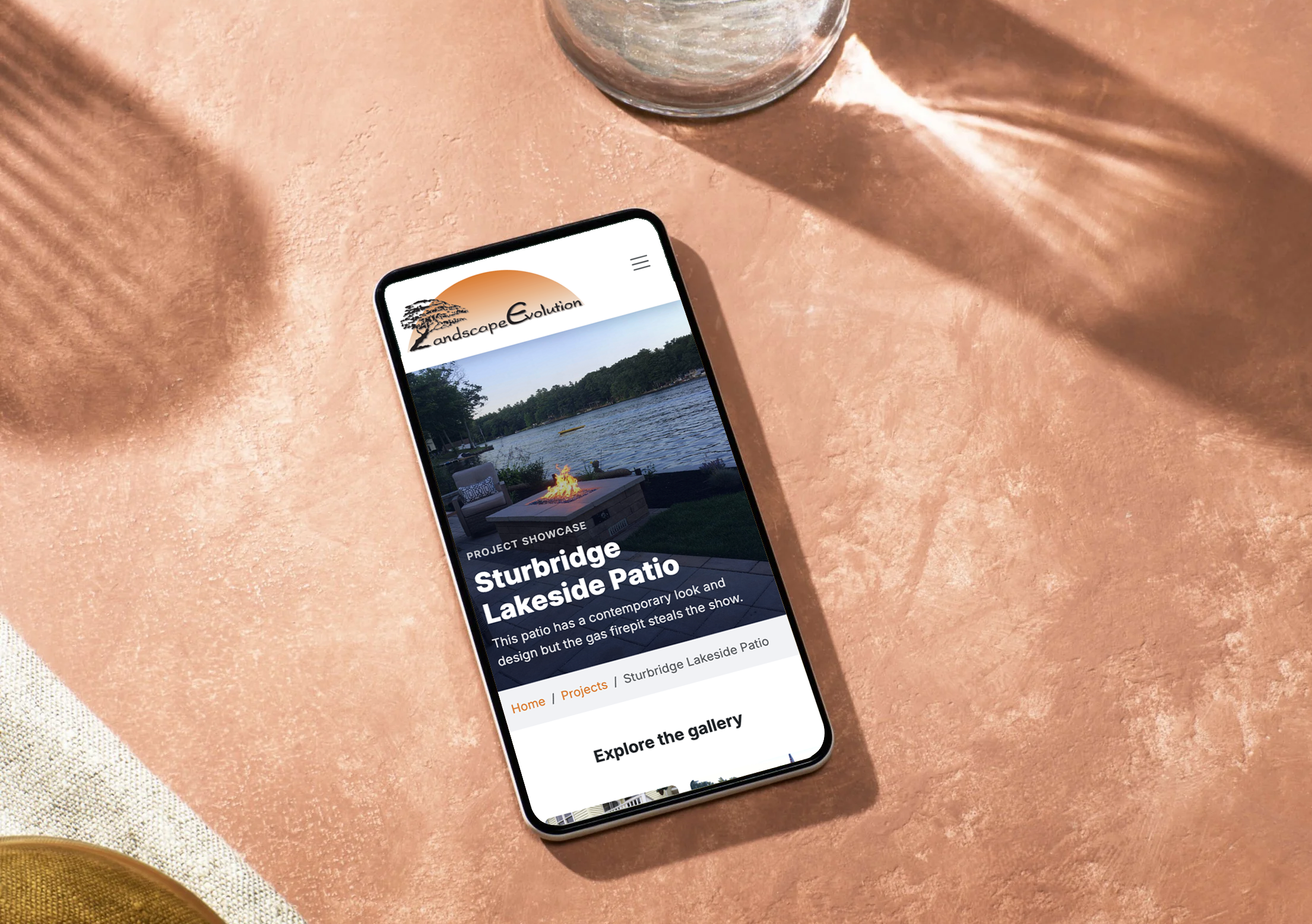 Smartphone on peach-colored surface displaying a webpage about Sturbridge Lakeside Patio project, with a glass of water nearby.