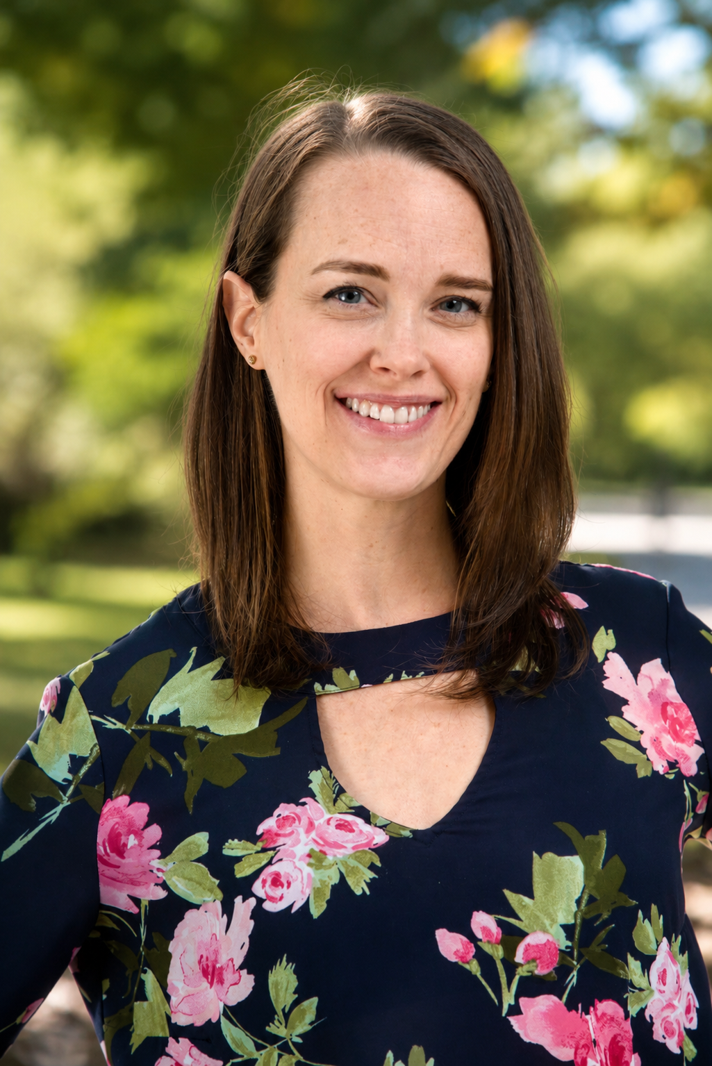 A woman with shoulder-length brown hair in a park with green trees, smiling at the camera, wearing a dark floral blouse with pink and green flowers.