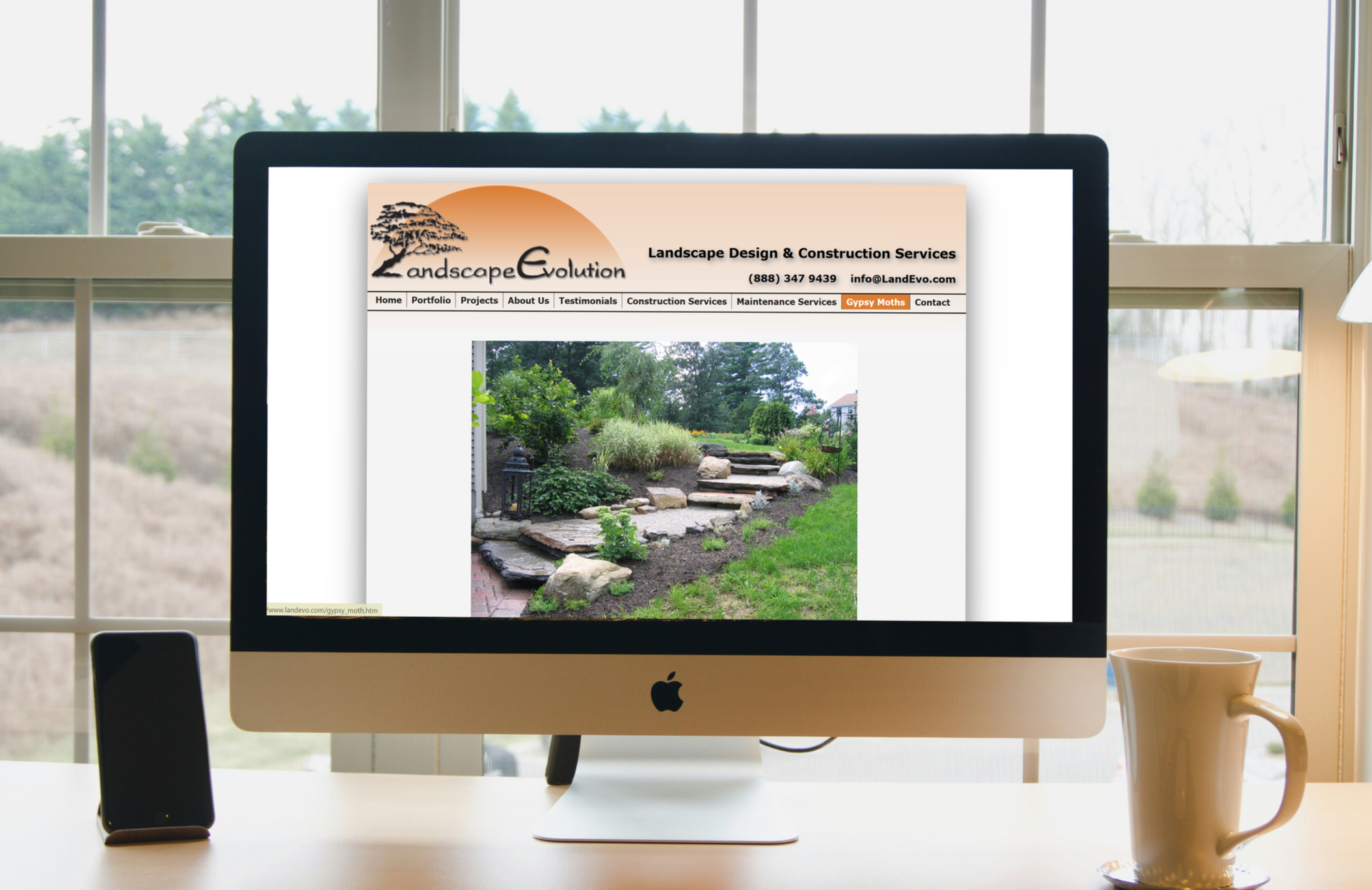 An iMac computer on a white desk displaying a landscape gardening website. The background shows a window with trees and outdoor scenery.