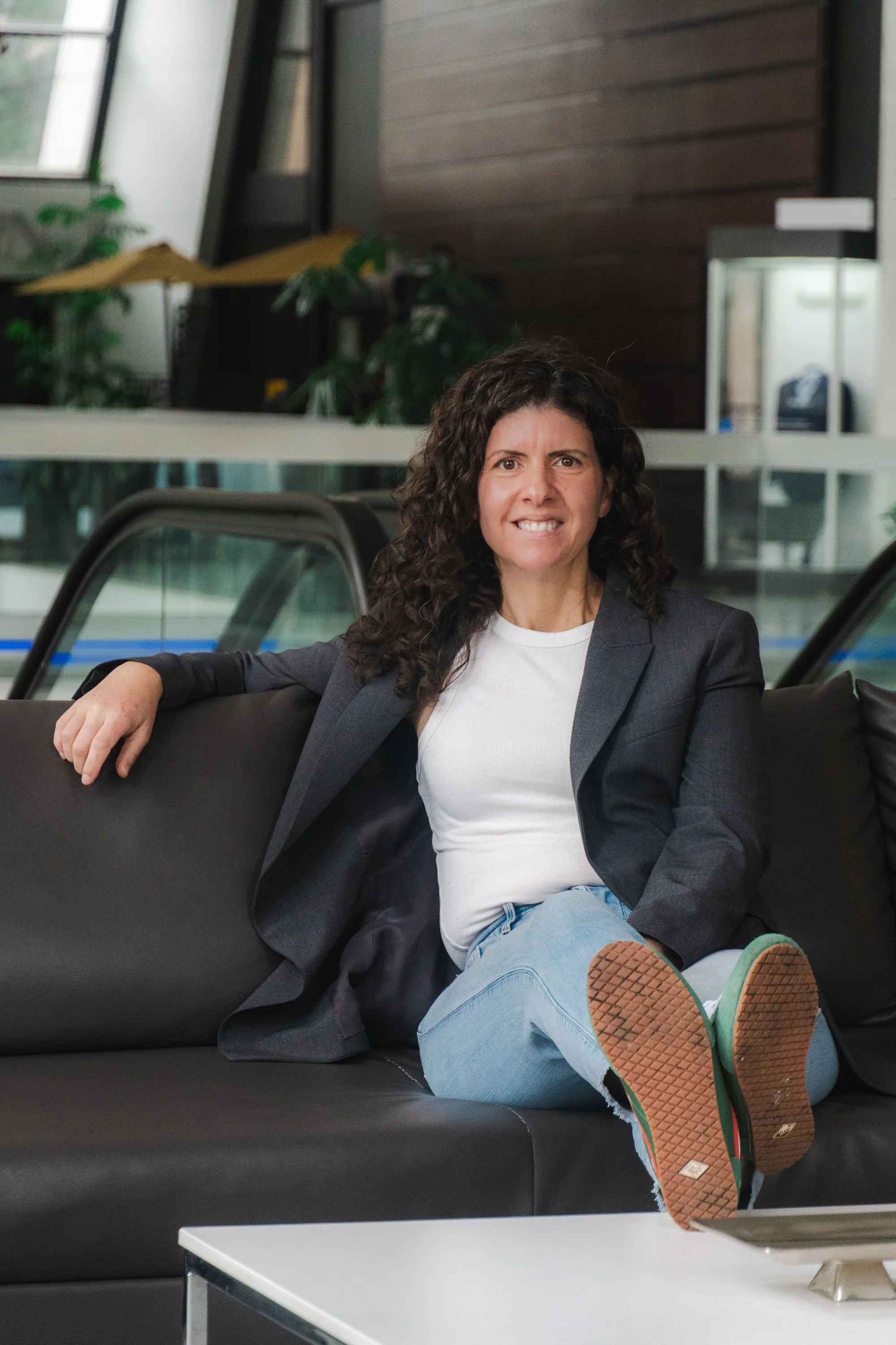 A woman sitting on a black couch in an indoor space, wearing a blazer, white shirt, and jeans, with her legs crossed and feet up, showing sneakers on display.