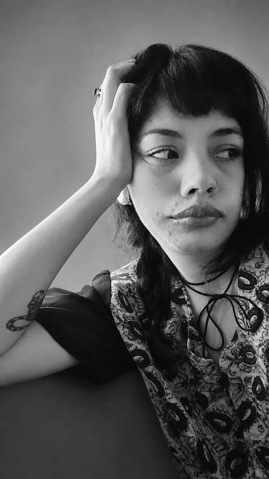 A woman with short dark hair and tattoos, wearing a patterned shirt, resting her head on her hand, and looking to the side, in black and white.