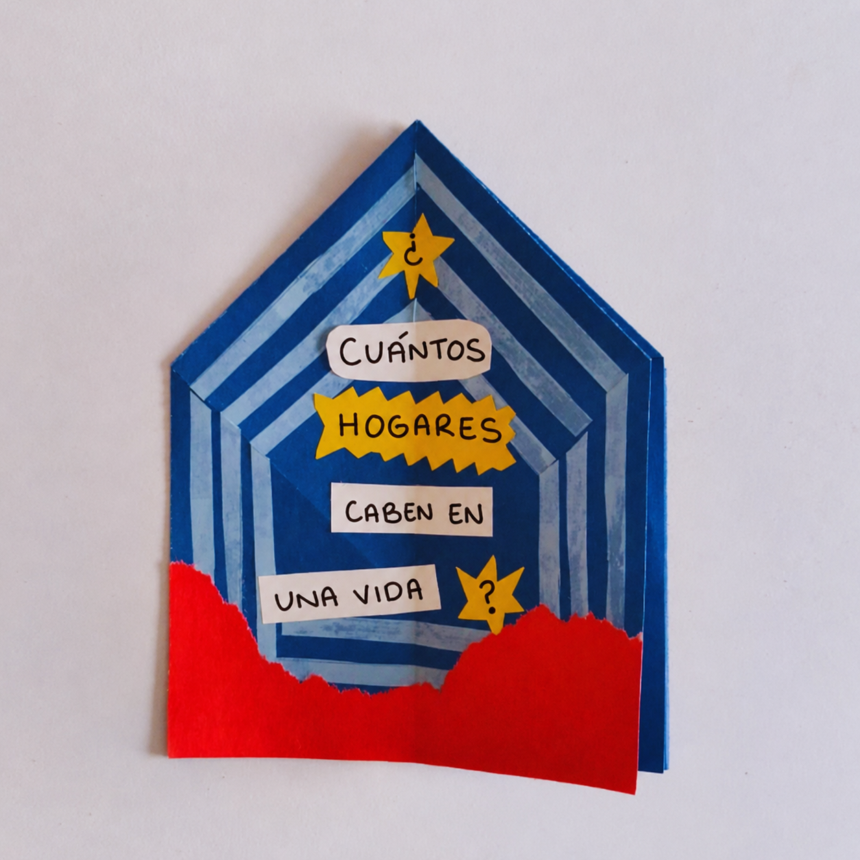 A colorful paper craft with a blue striped house shape and red grass at the bottom. It has Spanish text inside: "¿Cuántos hogares caben en una vida?" with some words highlighted in yellow and a yellow star with a question mark.