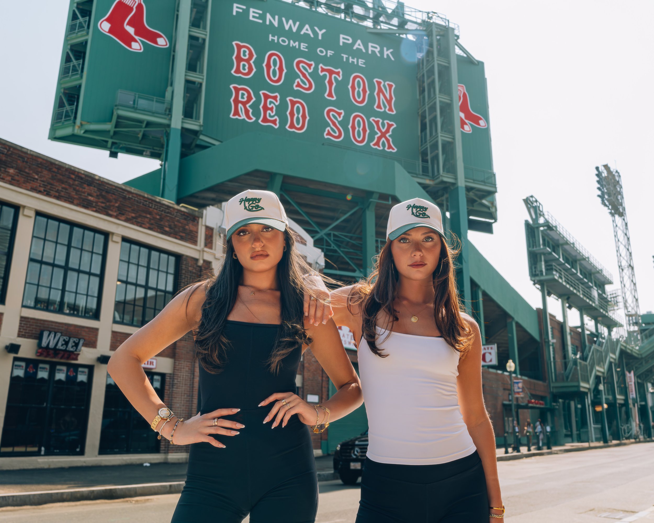 Two young women standing close together in front of Fenway Park in Boston, wearing white and black tops and baseball caps, with the Fenway Park sign visible in the background.