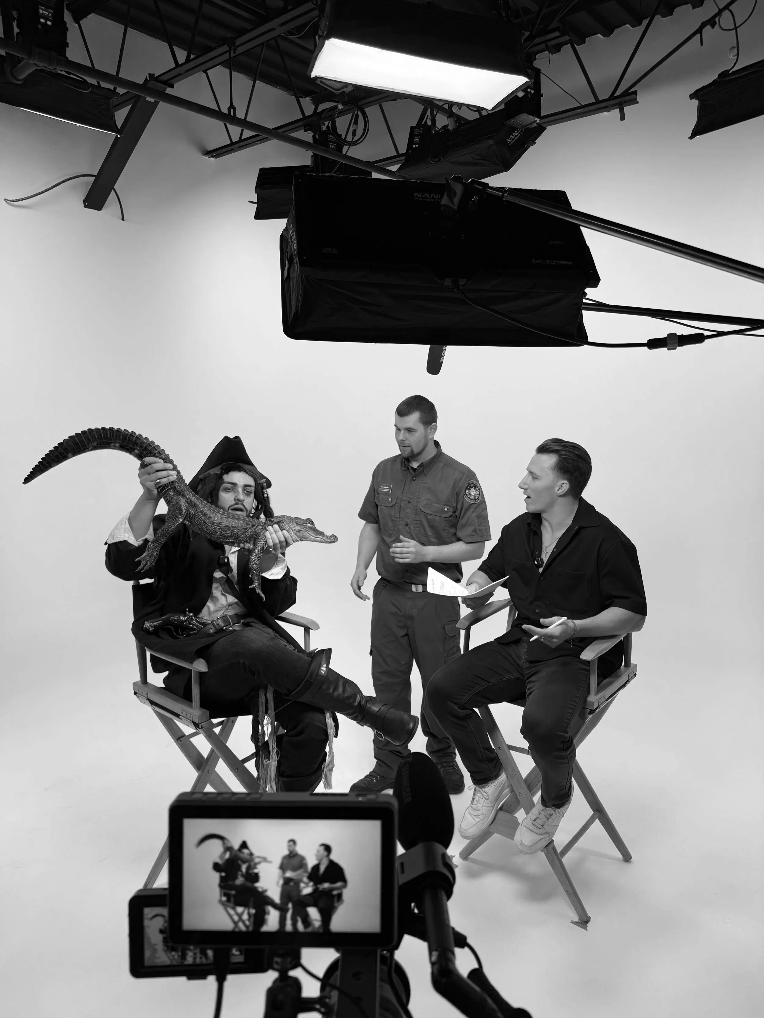 A behind-the-scenes photo of a video shoot or interview. Three men are in a studio with a plain light-colored background; one is holding a crocodile, and the other two are engaged in conversation. Studio lighting equipment and a camera monitor are visible.