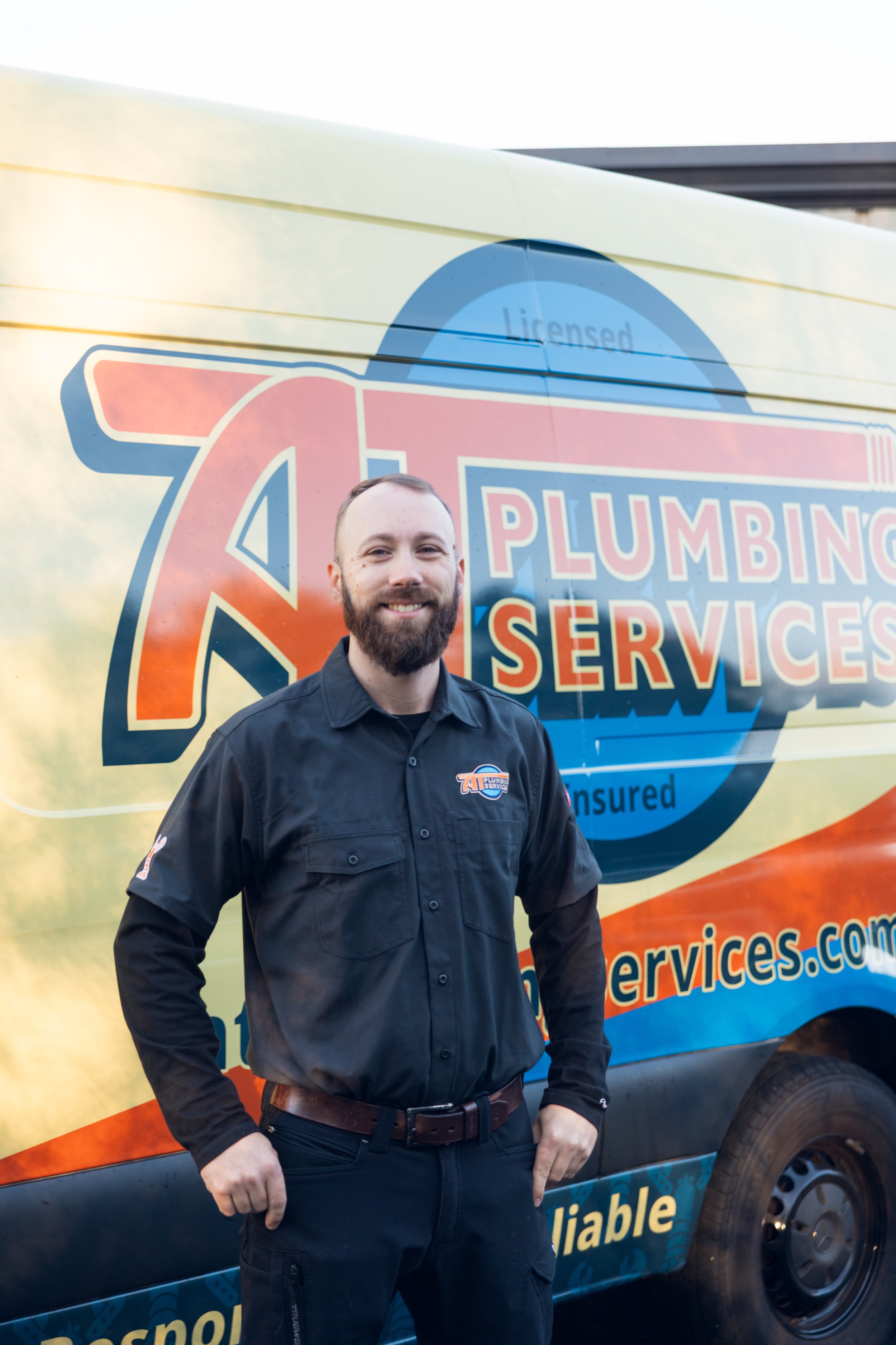 A smiling man with a beard wearing a black shirt standing in front of a yellow and blue service van that reads 'Plumbing Services'.