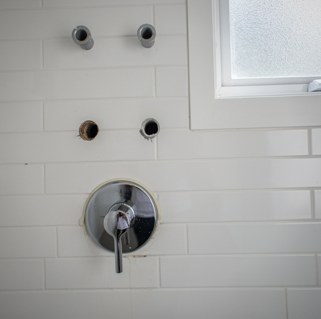 White subway tile wall with four holes and a circular valve fixture with handle below them, next to a window.