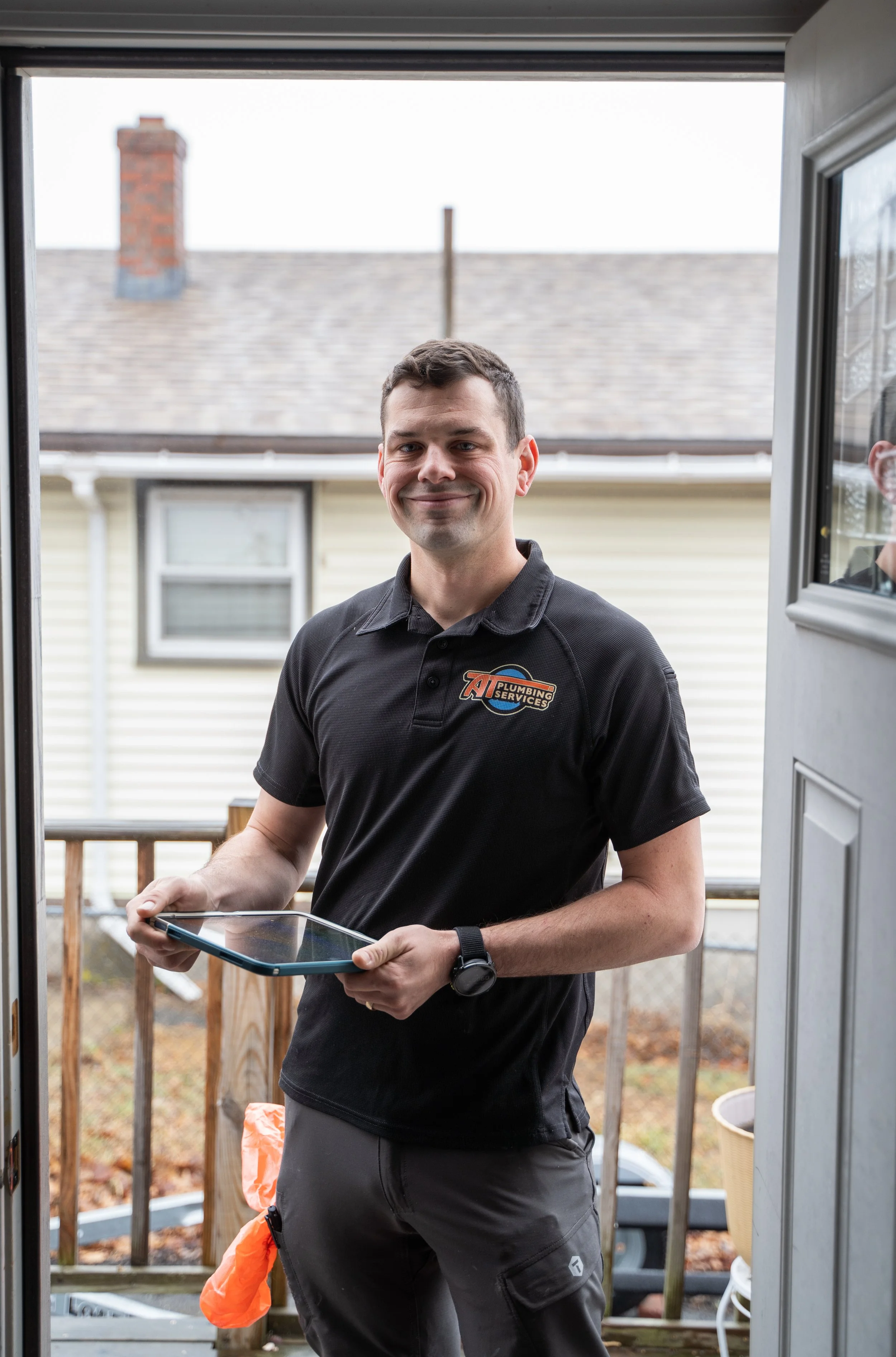 A man stands at a doorway, smiling, holding a tablet device. He is wearing a black polo shirt with a logo and gray pants. Behind him is a residential backyard with a wooden fence, an overcast sky, and part of a house with beige siding visible.