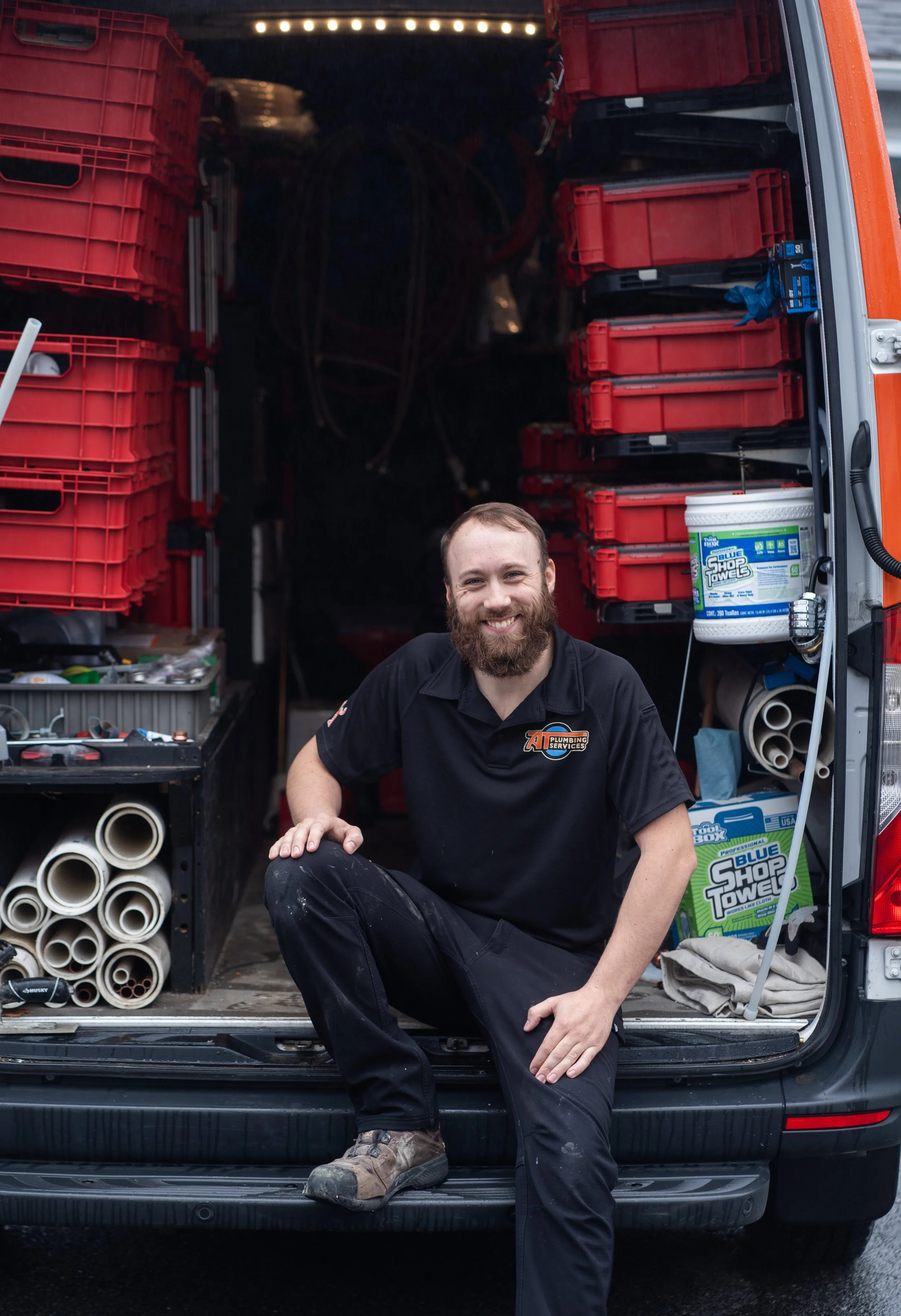 A smiling man with a beard sitting on the back of a truck filled with plumbing supplies, including red toolboxes, pipes, and cleaning products.