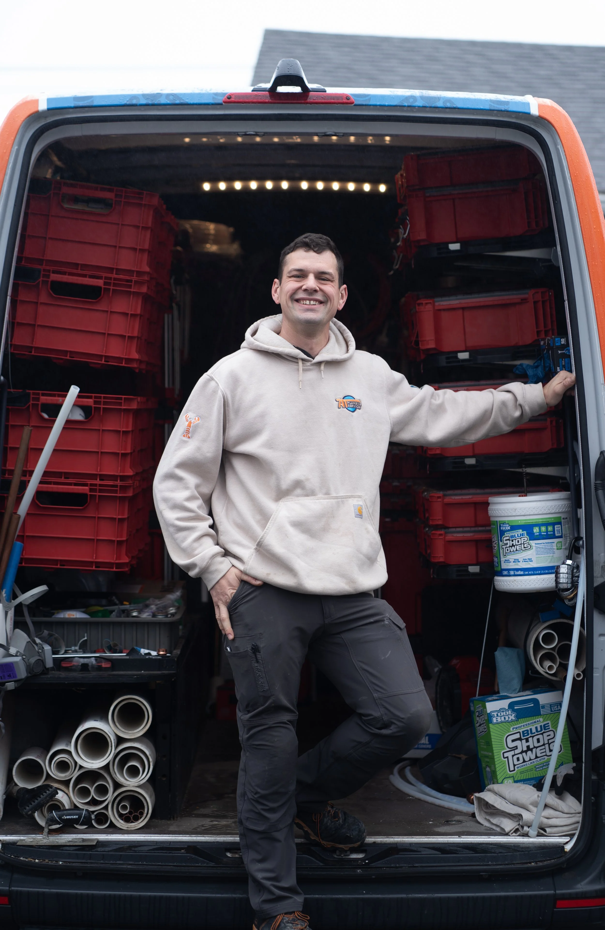 A man smiling and standing in the open back of a truck filled with red plastic storage bins, tools, and supplies.