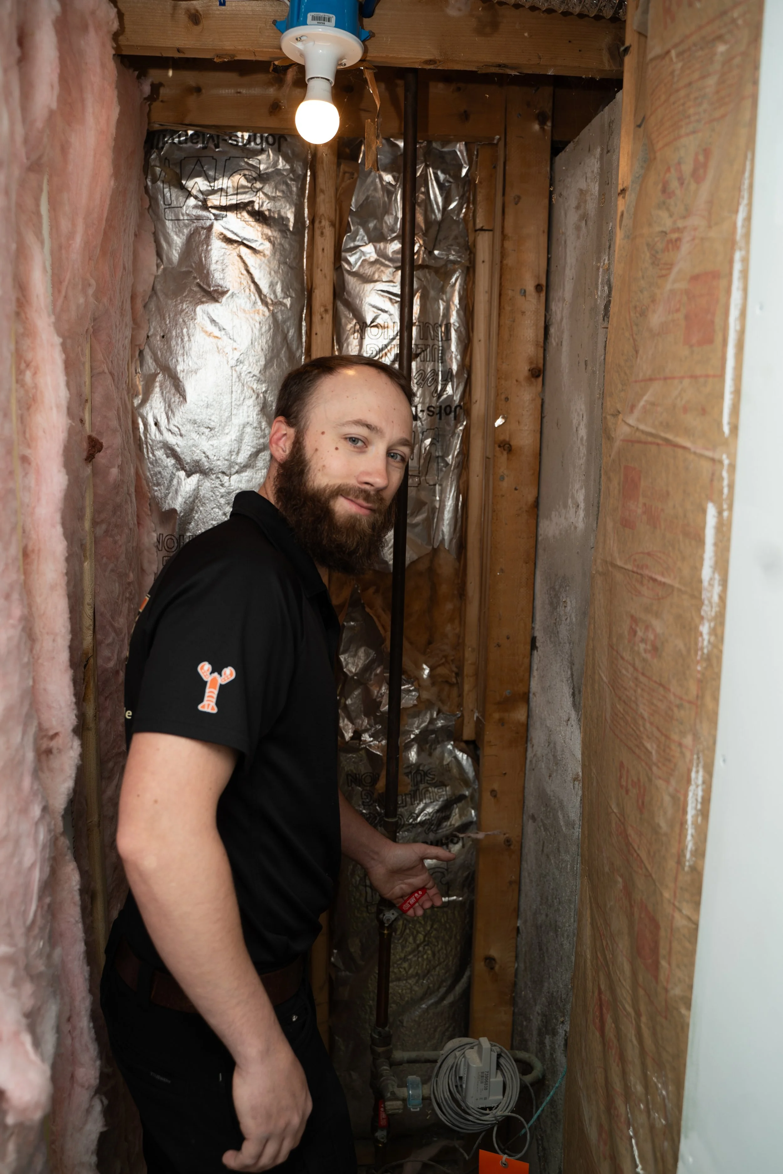 A man with a beard and short hair standing in a small, unfinished room with pink insulation on the left wall, metallic insulation behind him, and wooden framing. He is holding a tool near plumbing pipes at the bottom of the image and looking at the camera. A lit lightbulb hangs from the ceiling.