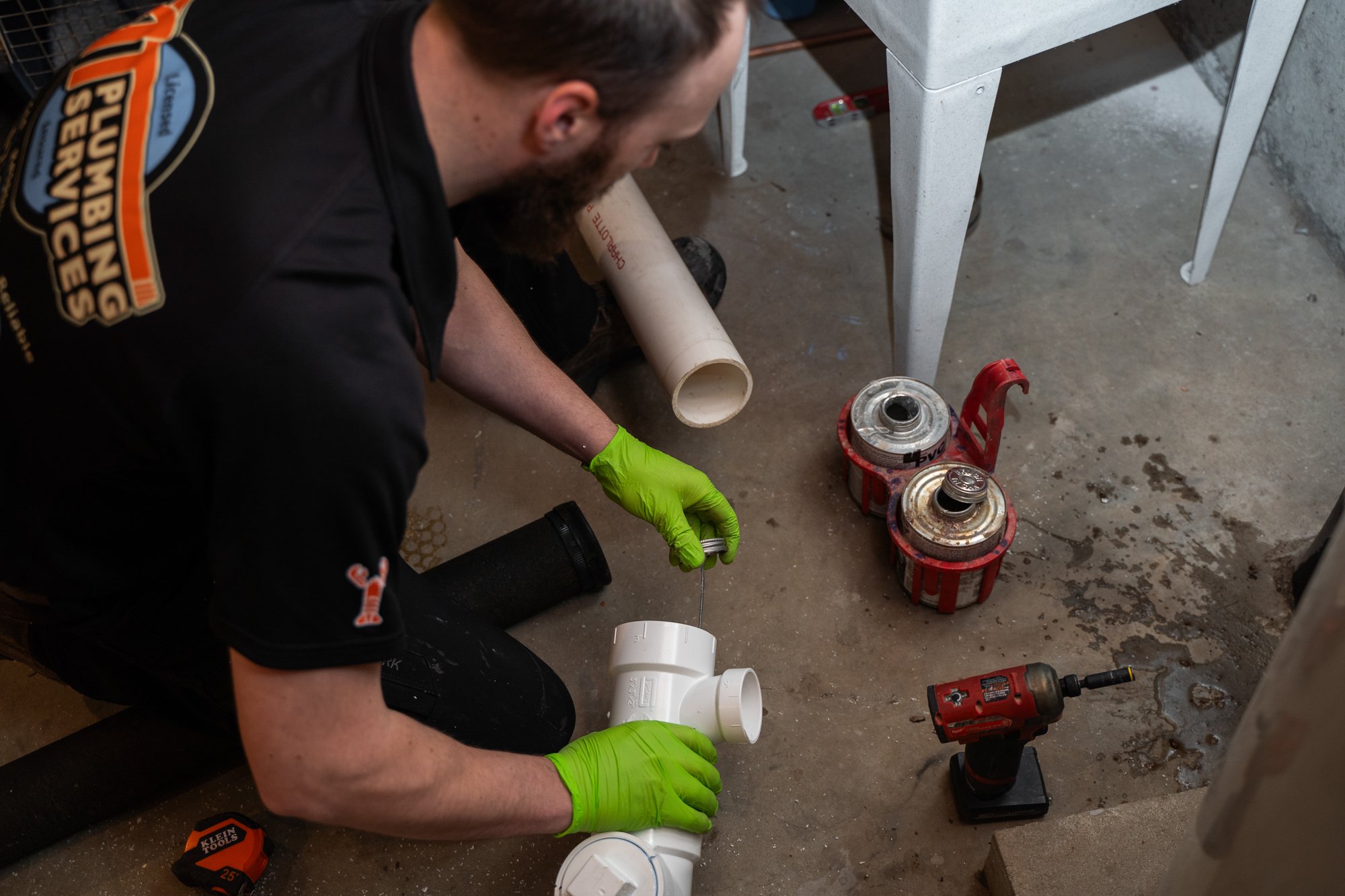 A man wearing a black shirt with orange and blue patches and bright green gloves is assembling white PVC pipes under a table. Various tools, including a drill and paint cans, are on the concrete floor around him.