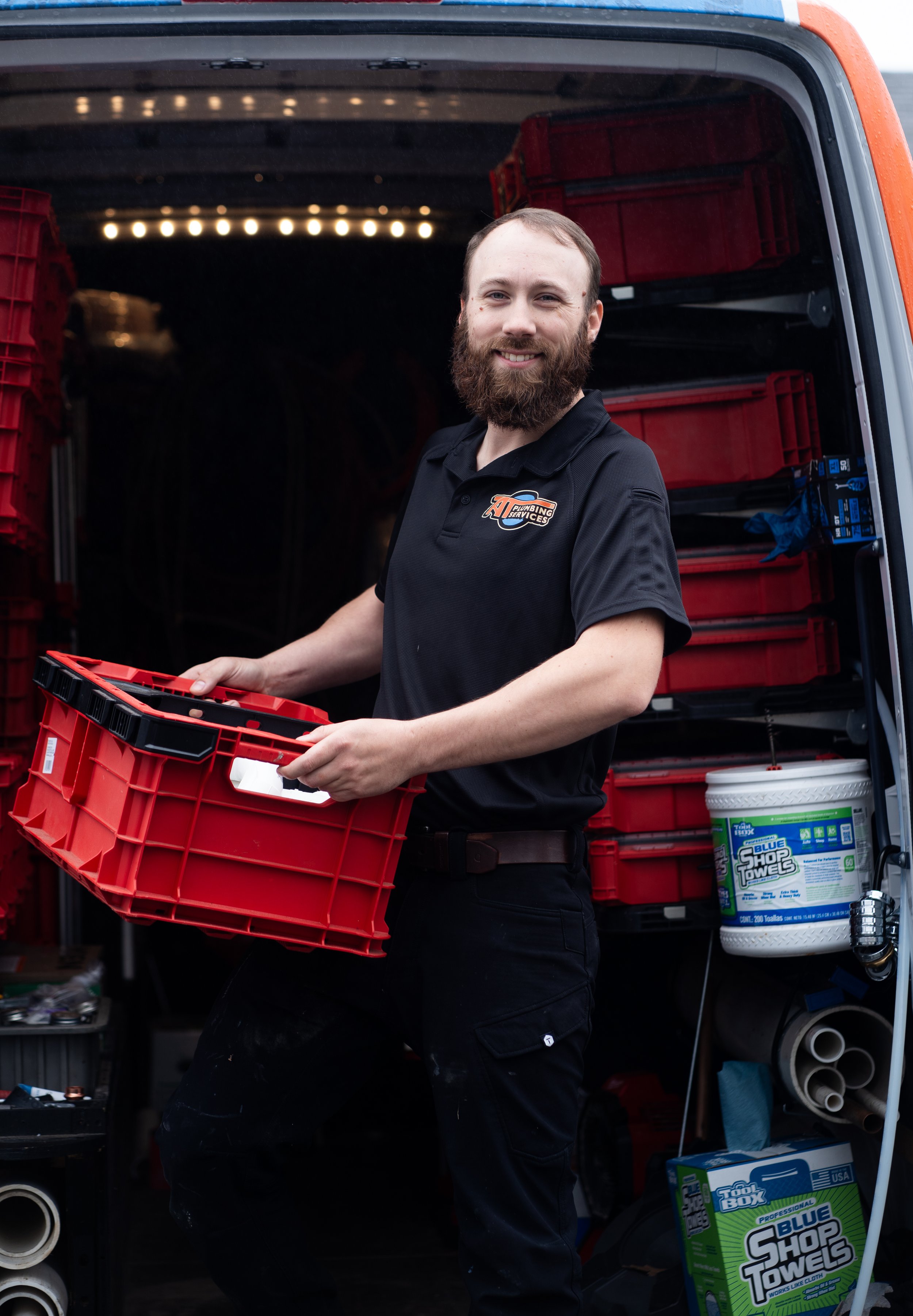 A man with a beard and a black uniform holding a red plastic crate inside a vehicle with shelves filled with red crates and cleaning supplies, including a large container of blue shop towels.