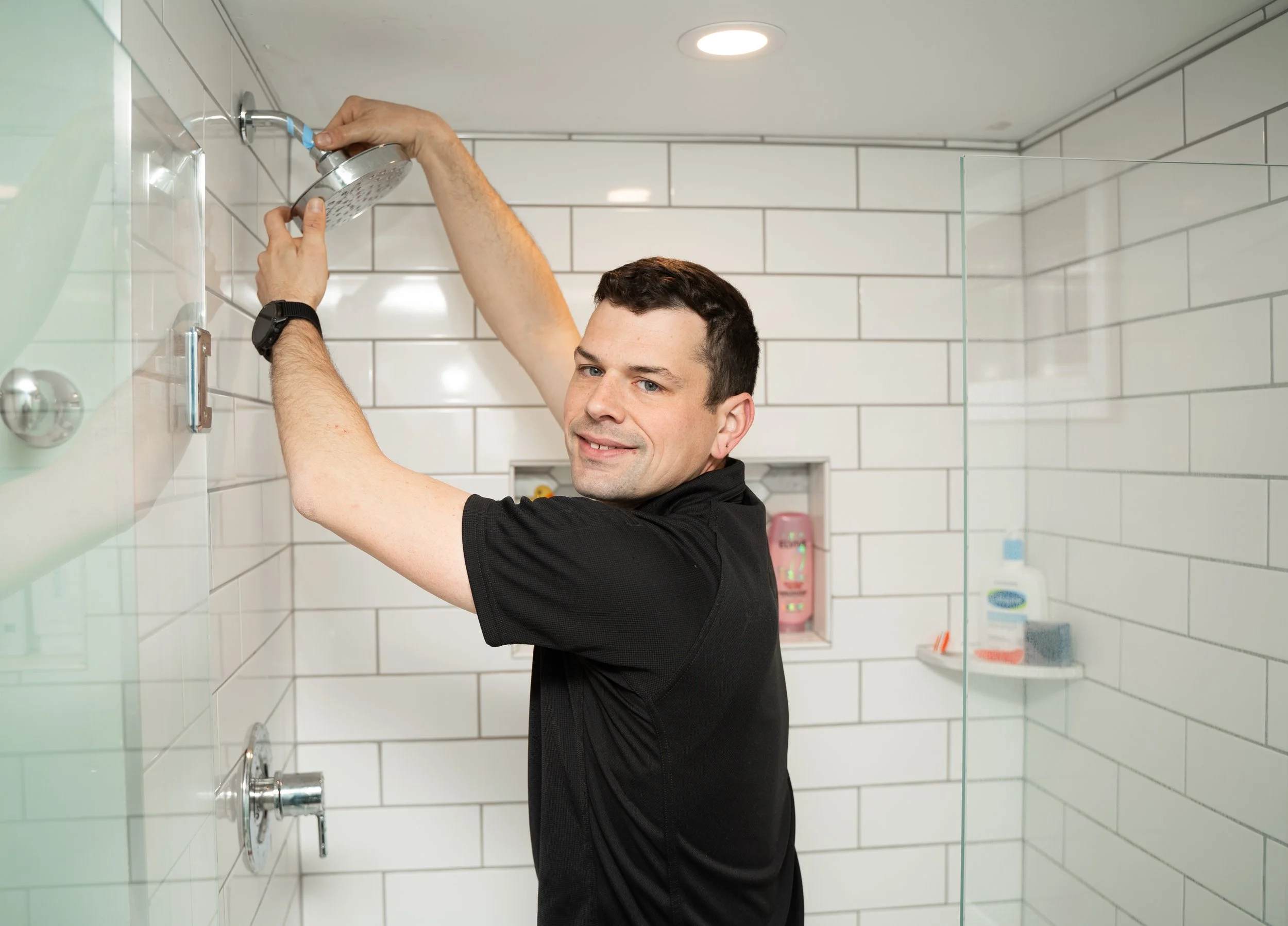 A man with short dark hair in a black shirt is cleaning a showerhead in a bathroom with white tile walls, using both hands.