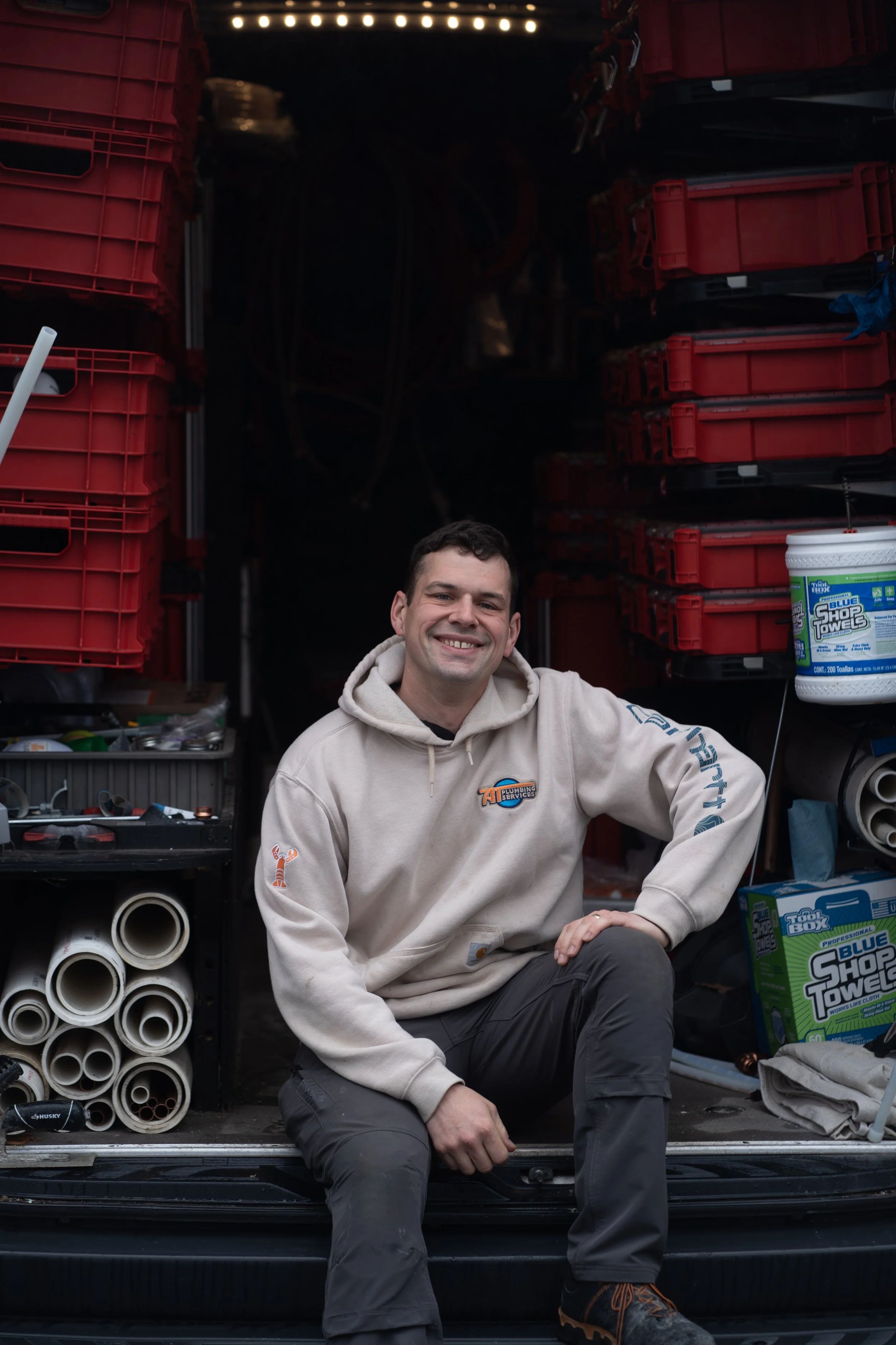 A young man smiling and sitting in the back of a truck with various tools, pipes, and storage containers around him.