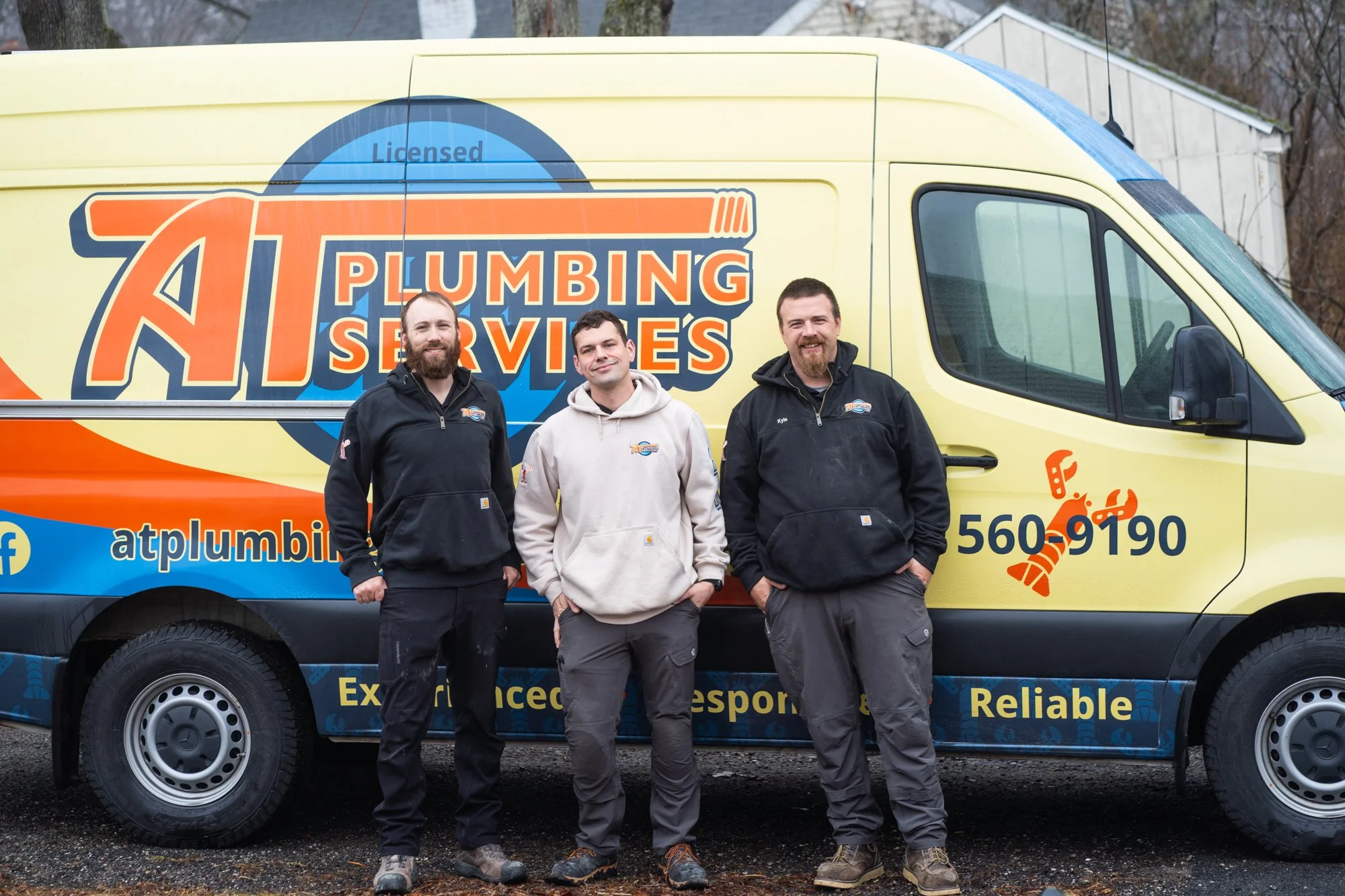 Three men standing in front of a yellow plumbing service van with the company logo and contact information, outdoors on a cloudy day.