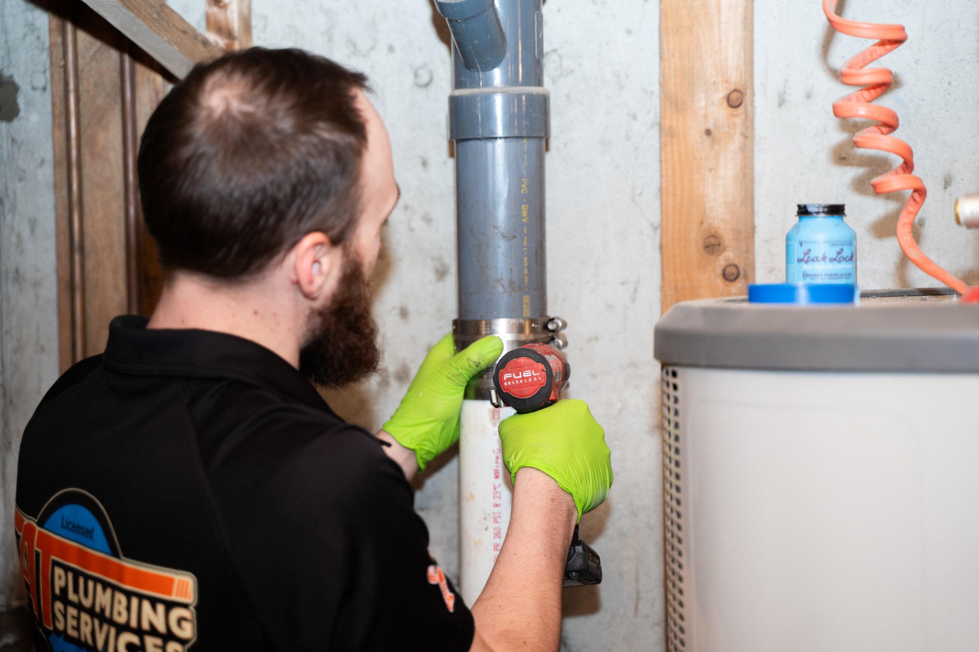 A man with brown hair and a beard wearing green gloves and a black shirt with plumbing company logo, working on a vertical pipe with a cordless drill in a basement or utility room.