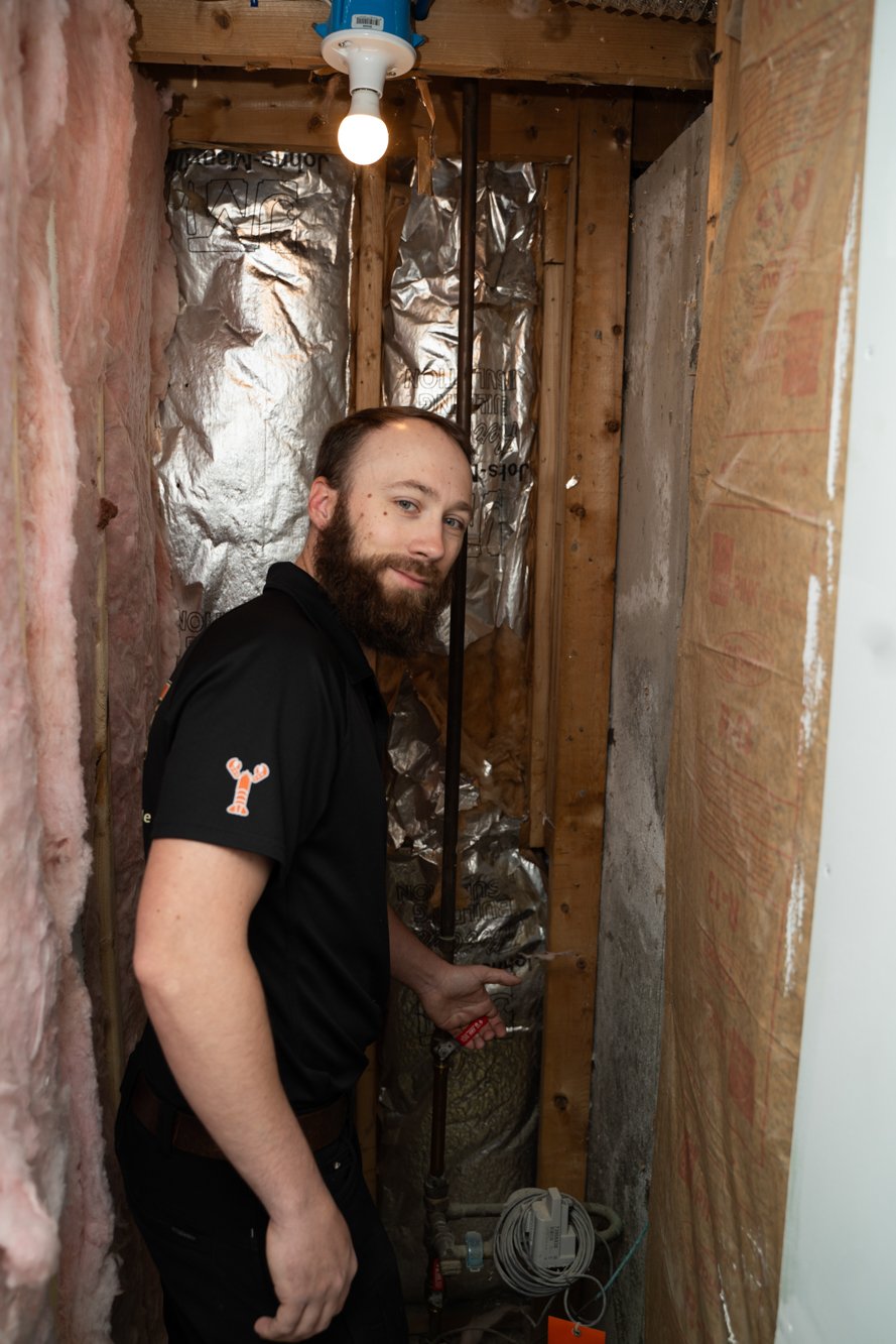 A man inspecting plumbing inside a wall cavity during a renovation, surrounded by insulation and wood framing.