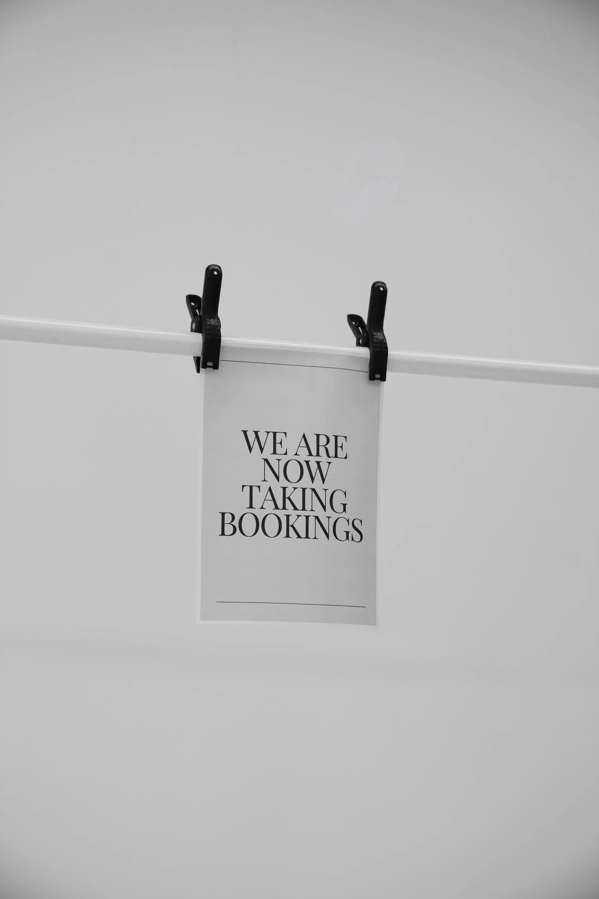 A printed sign with the message "We are now taking bookings" hangs from a white horizontal rod using black clips.