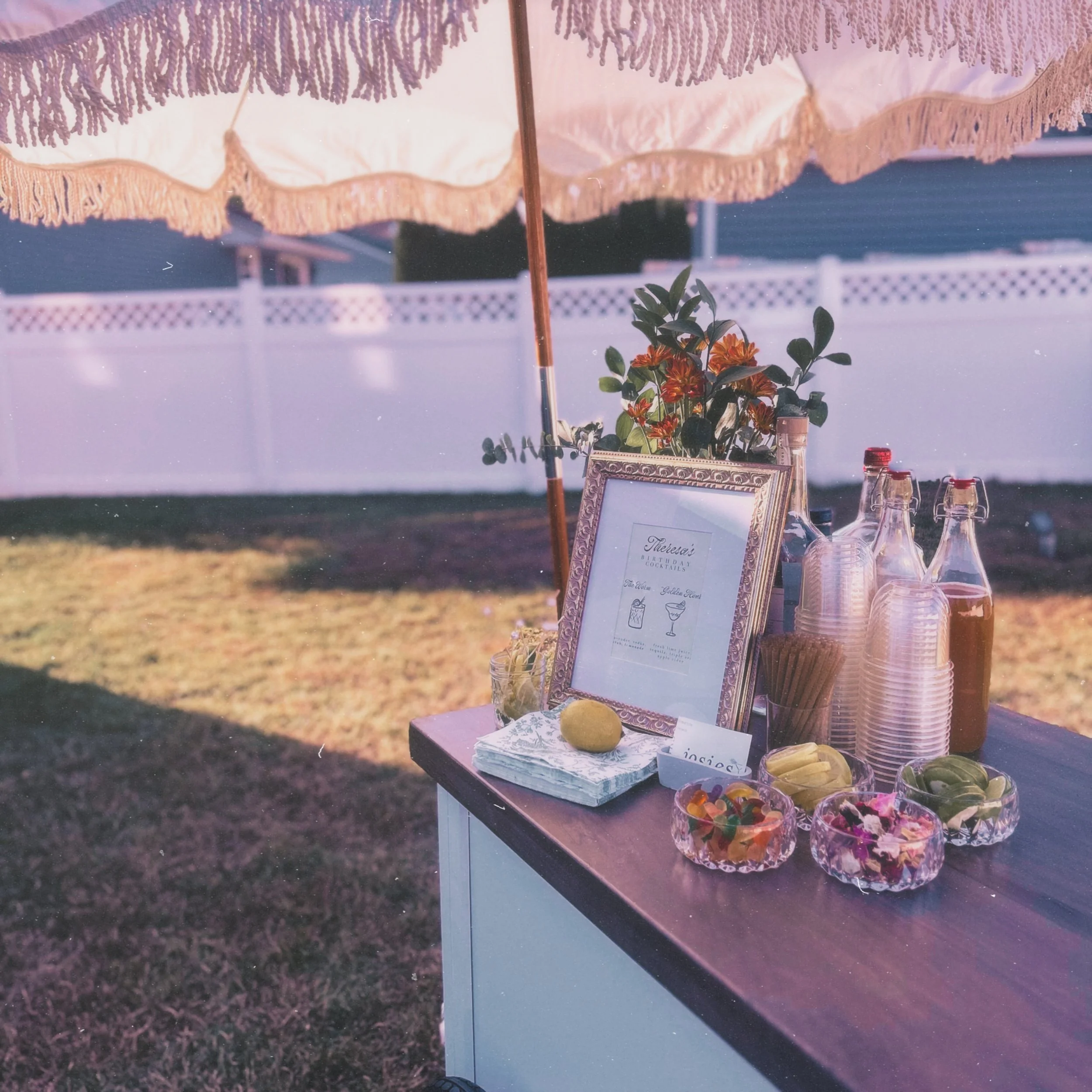 Outdoor table with pink and orange flowers, glass bottles, a framed sign with drink menu, and a pink umbrella providing shade during sunset.