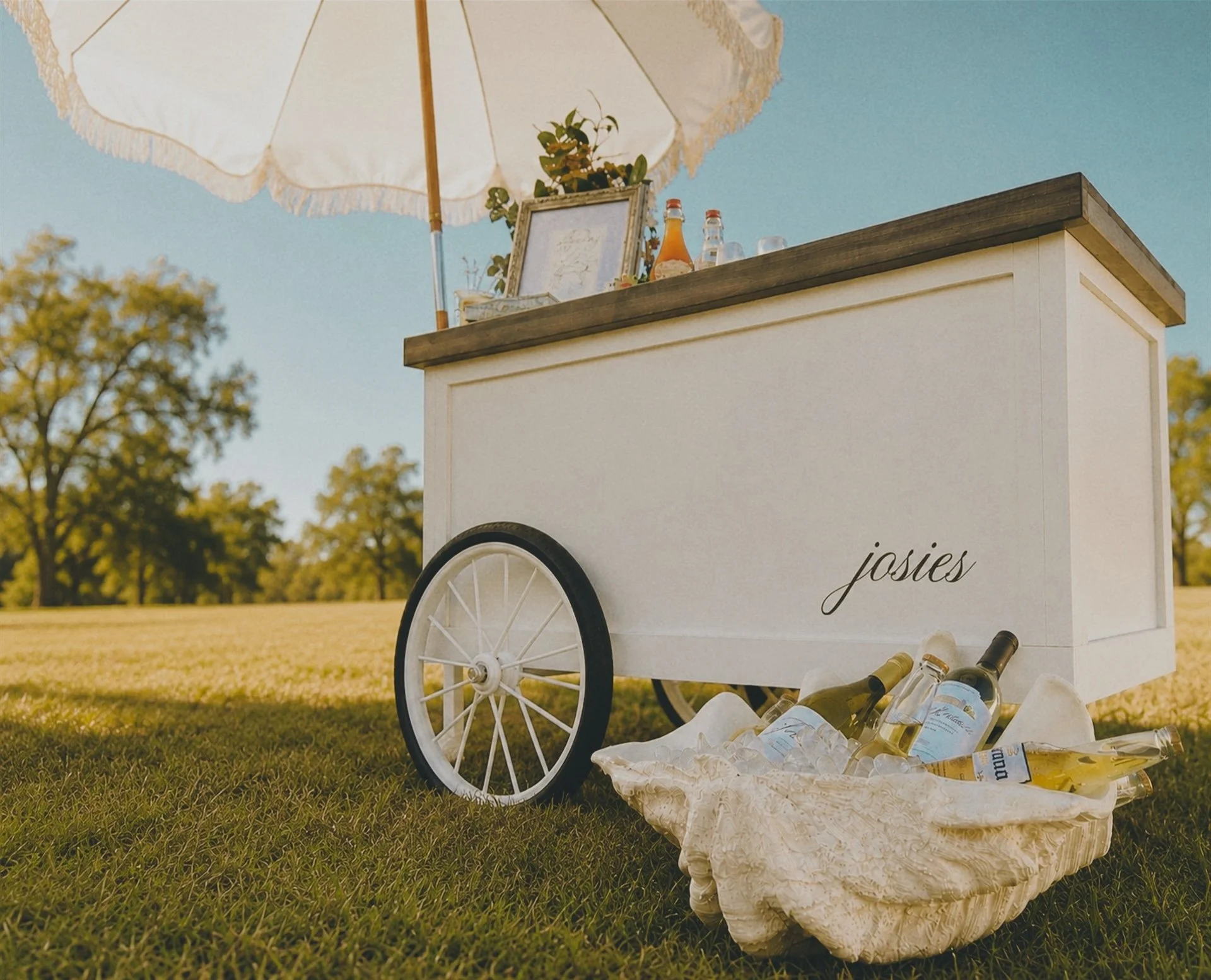 A white ice cream cart with the name 'Josies' written on it, situated outdoors on grass with trees in the background. The cart has a canopy umbrella, bottles of soda, a framed picture, and a potted plant on top. Los Angeles, California.