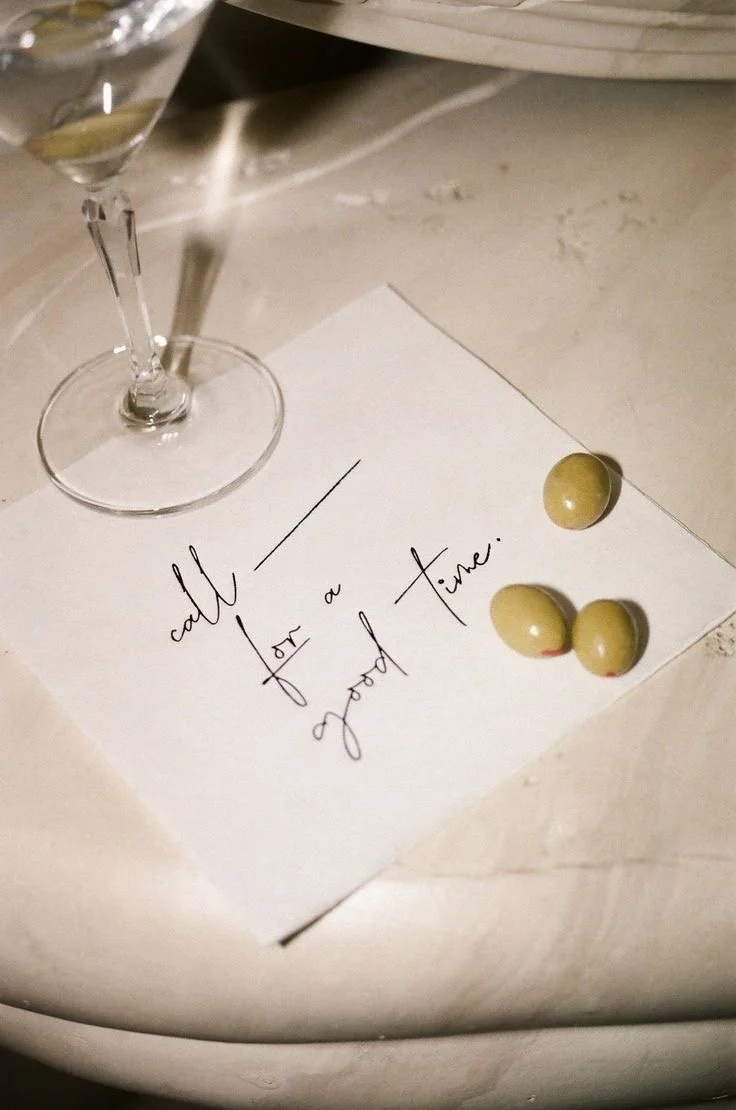 A glass of white wine, a handwritten note on white paper that says "call for a good time," and three green olives on the paper, placed on a marble surface.