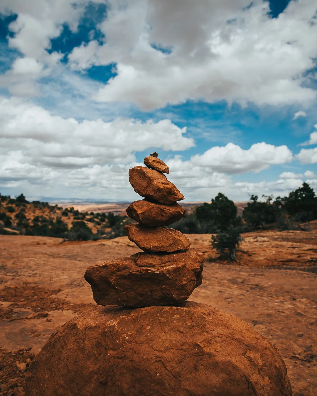 A balanced stack of five reddish-brown rocks on a large rock in a desert landscape with sparse vegetation and a cloudy blue sky.
