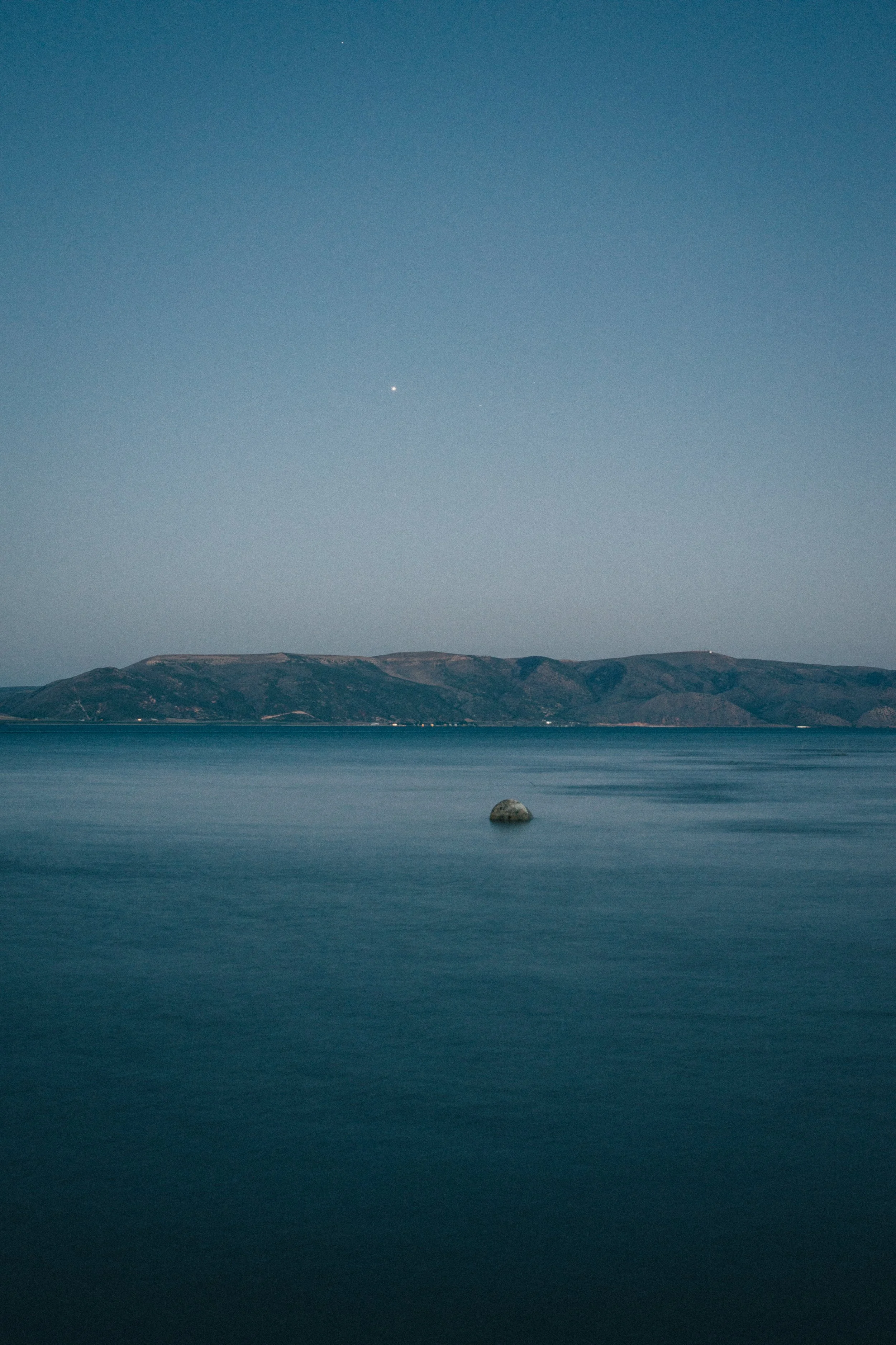 A calm body of water with a large rock in the foreground, a distant mountain range, and a clear sky with a bright planet or star visible