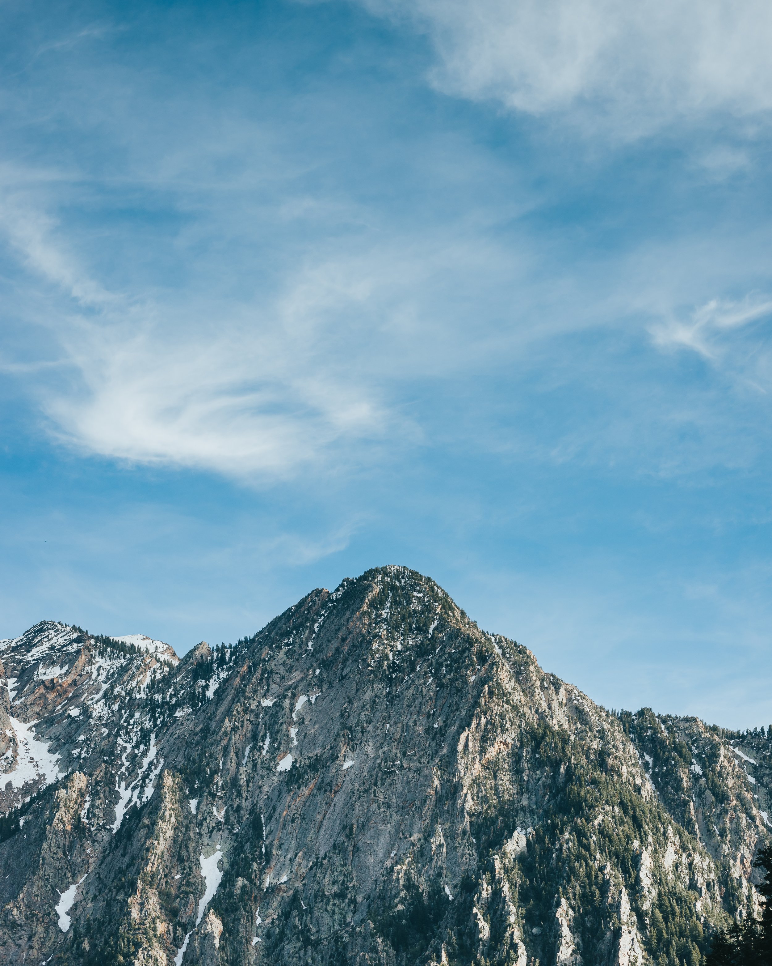 Snow-capped mountain with rocky face against a partly cloudy blue sky.