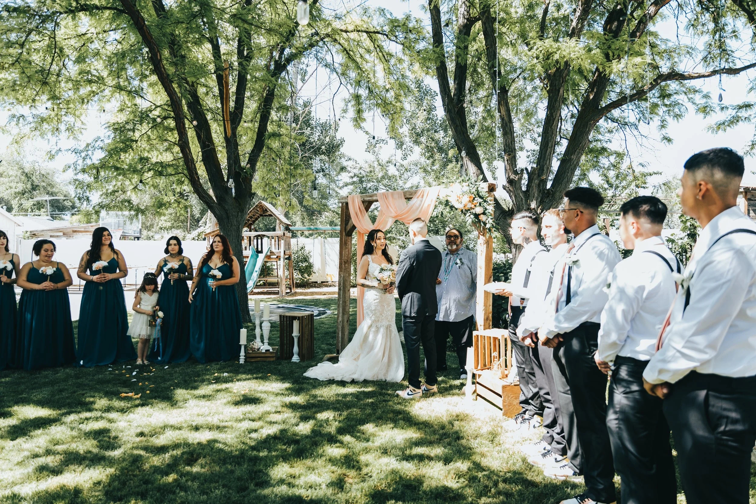 Outdoor wedding ceremony with a bride and groom, bridesmaids in navy dresses, and groomsmen in white shirts, set under tall trees with hanging lights and floral decorations.