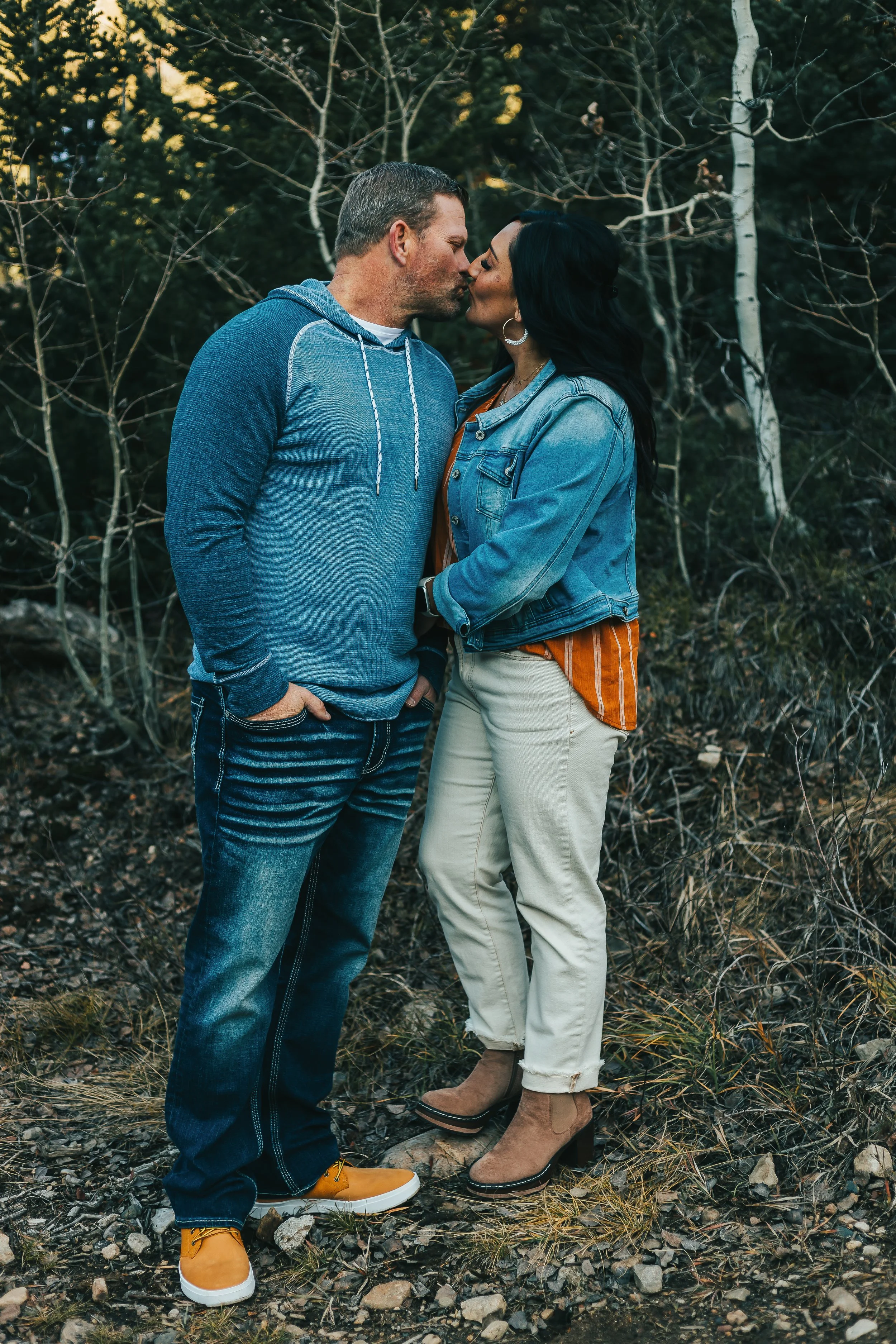 A couple sharing a kiss outdoors among trees with leafless branches, dressed casually with the man in a blue hoodie and jeans, and the woman in a denim jacket and light-colored pants, both wearing boots.