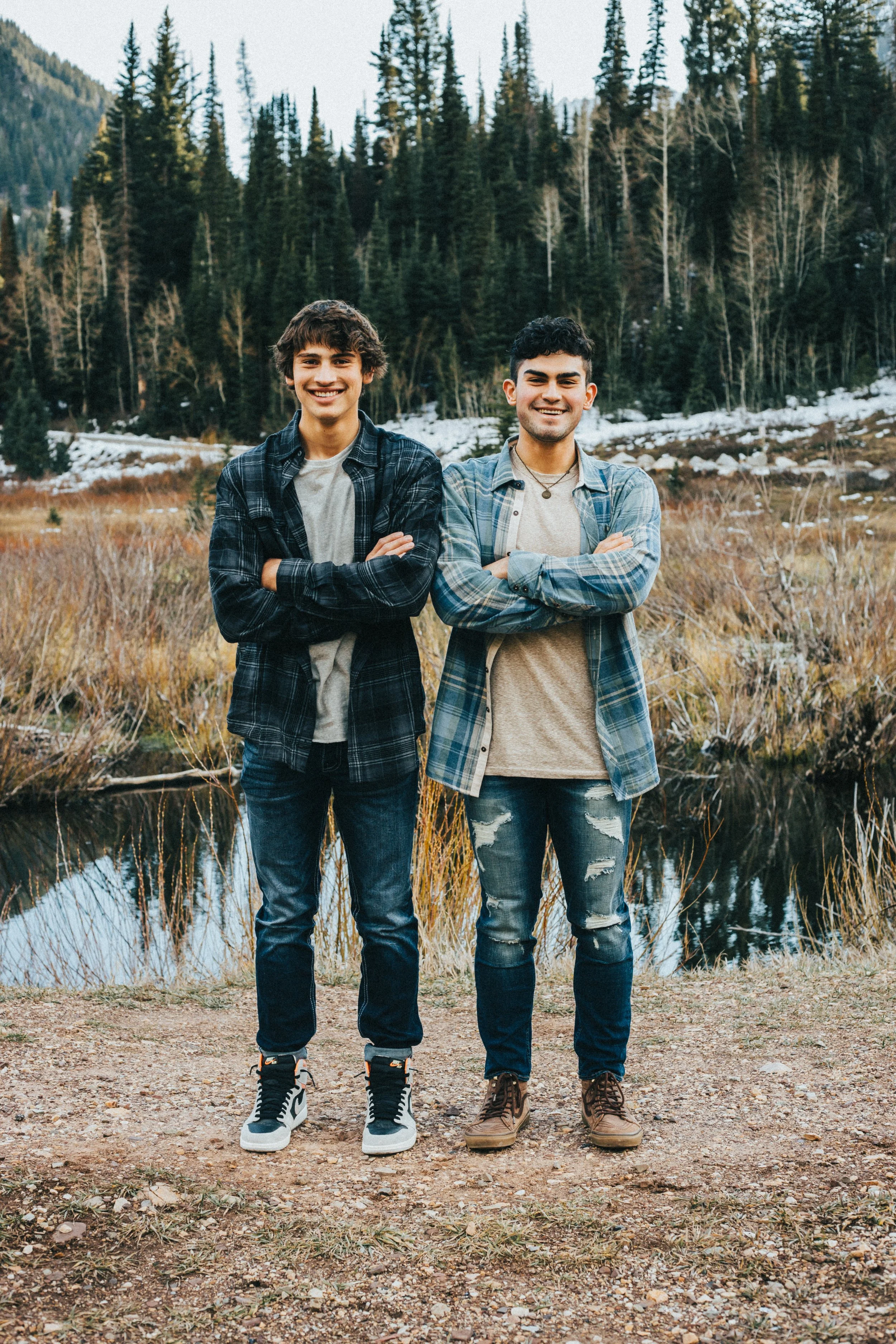 Two smiling young men standing outdoors with arms crossed in front of a forested mountain landscape with snow patches.